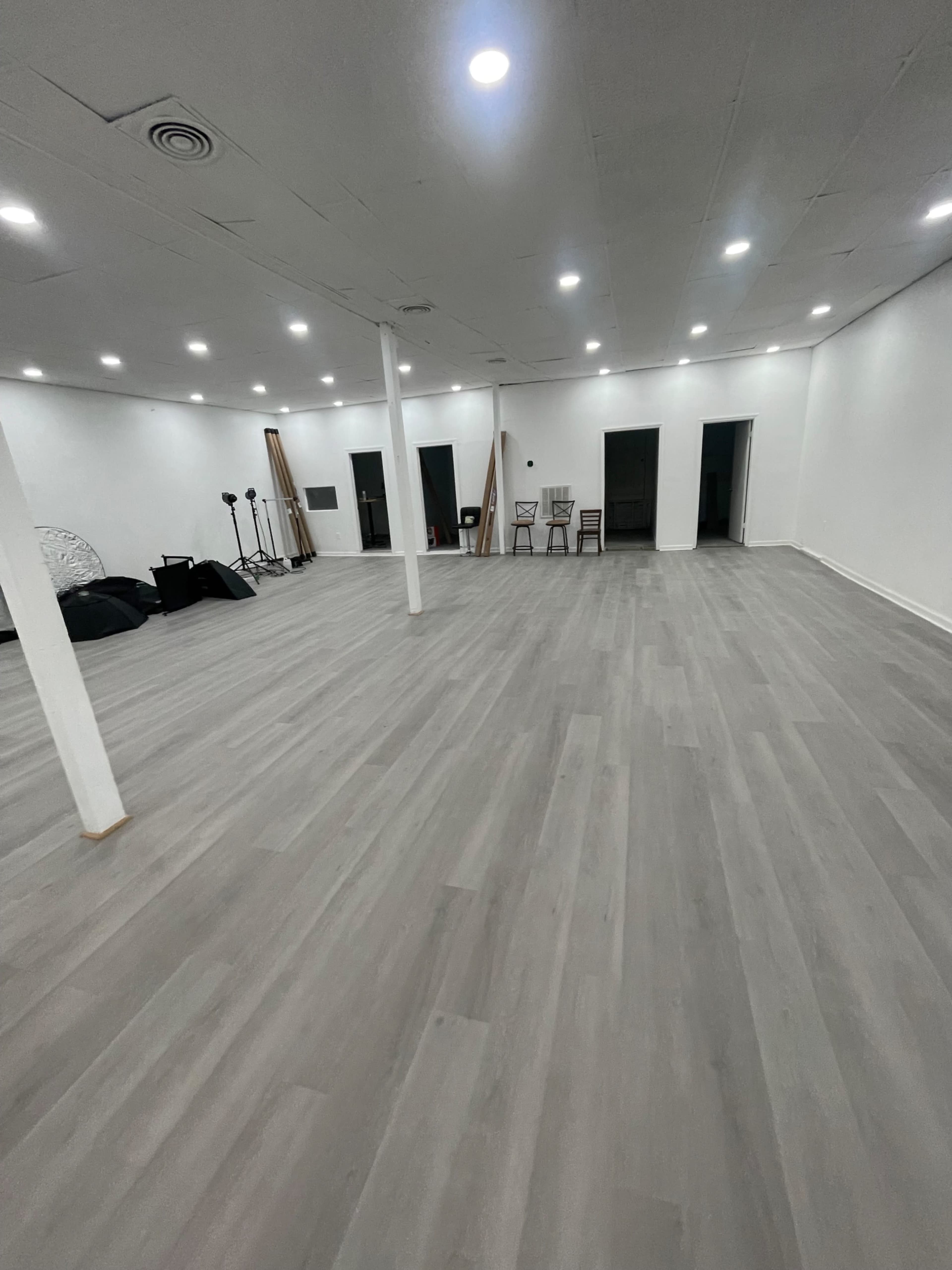 A spacious, empty room with gray wooden flooring, white walls, and several doors along one side.