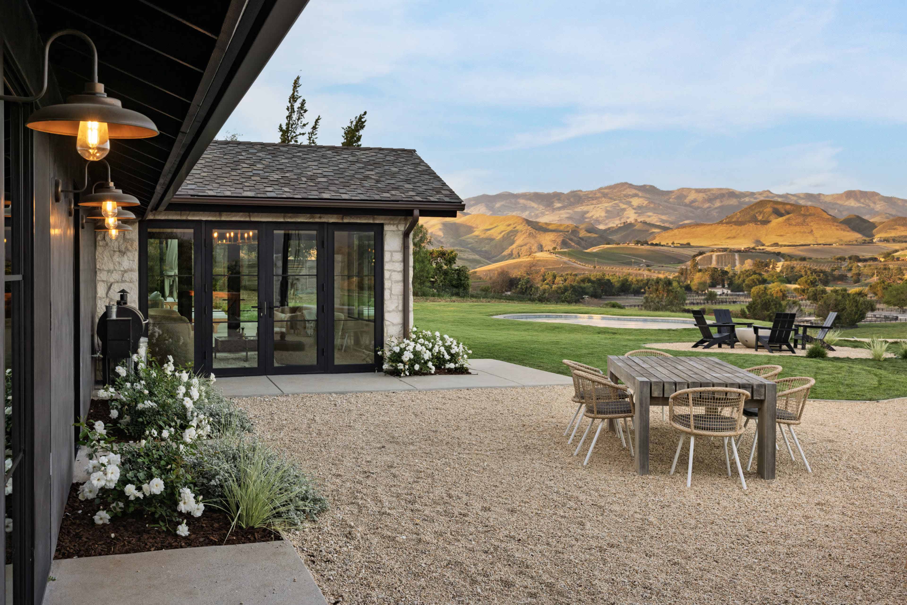 A modern patio features a circular dining table and chairs, with a view of mountains and green hills in the background.