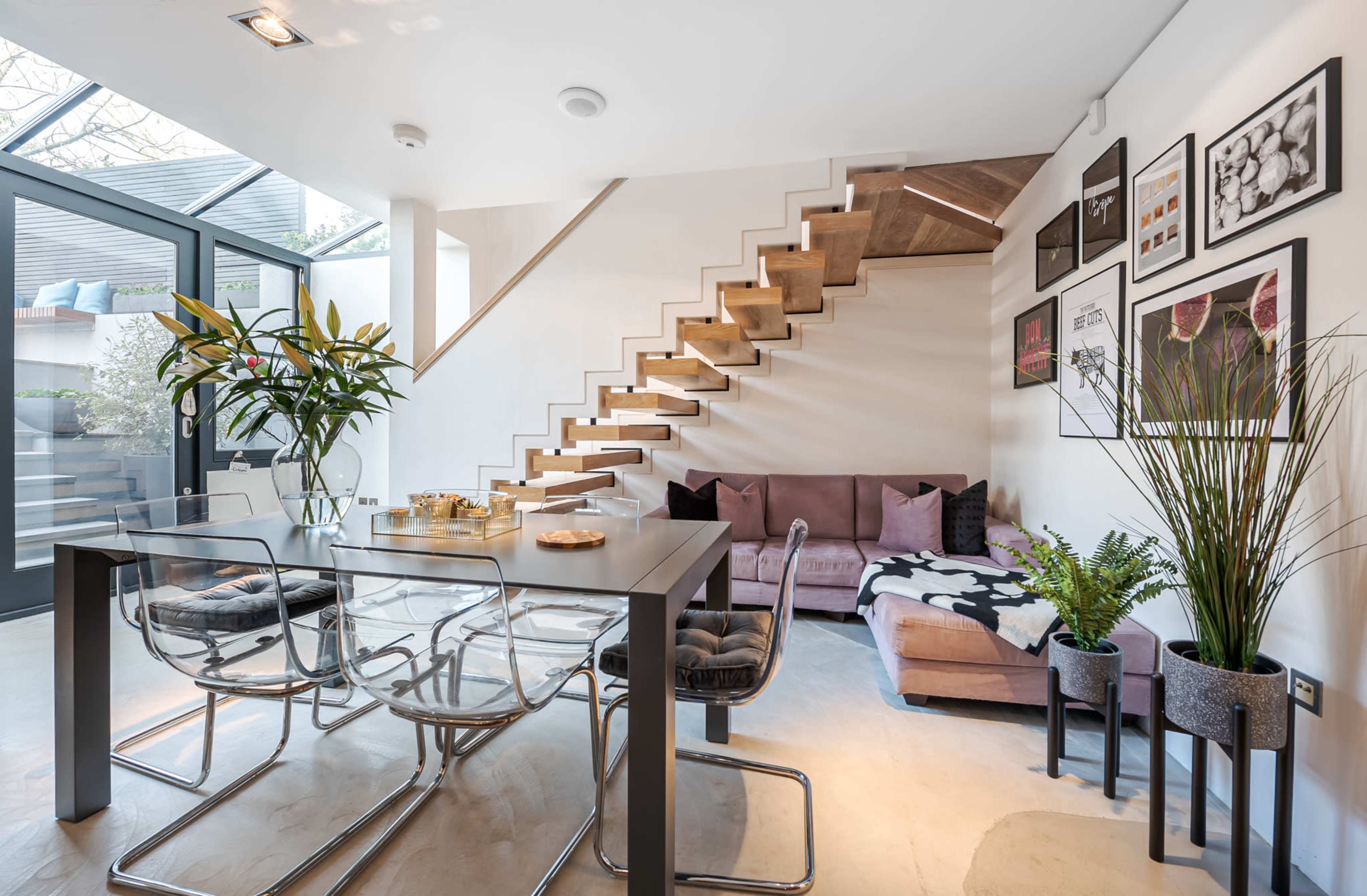 A modern dining area features a glass table with chrome chairs, a pink couch, and a staircase leading to an upper level, surrounded by potted plants and framed artwork on the walls.