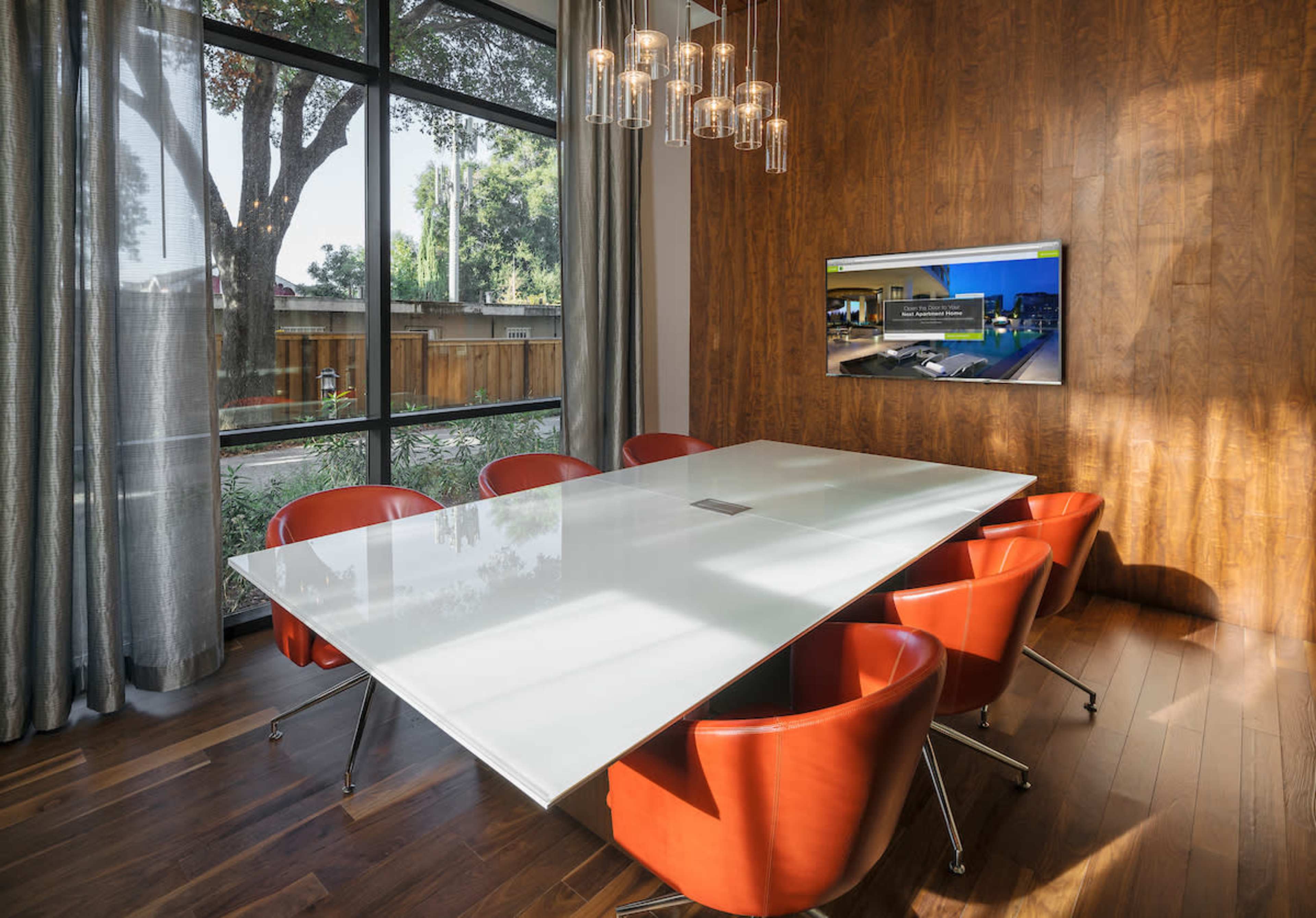 A modern conference room features a large white table surrounded by red chairs, with large windows overlooking a garden and a television mounted on the wall.