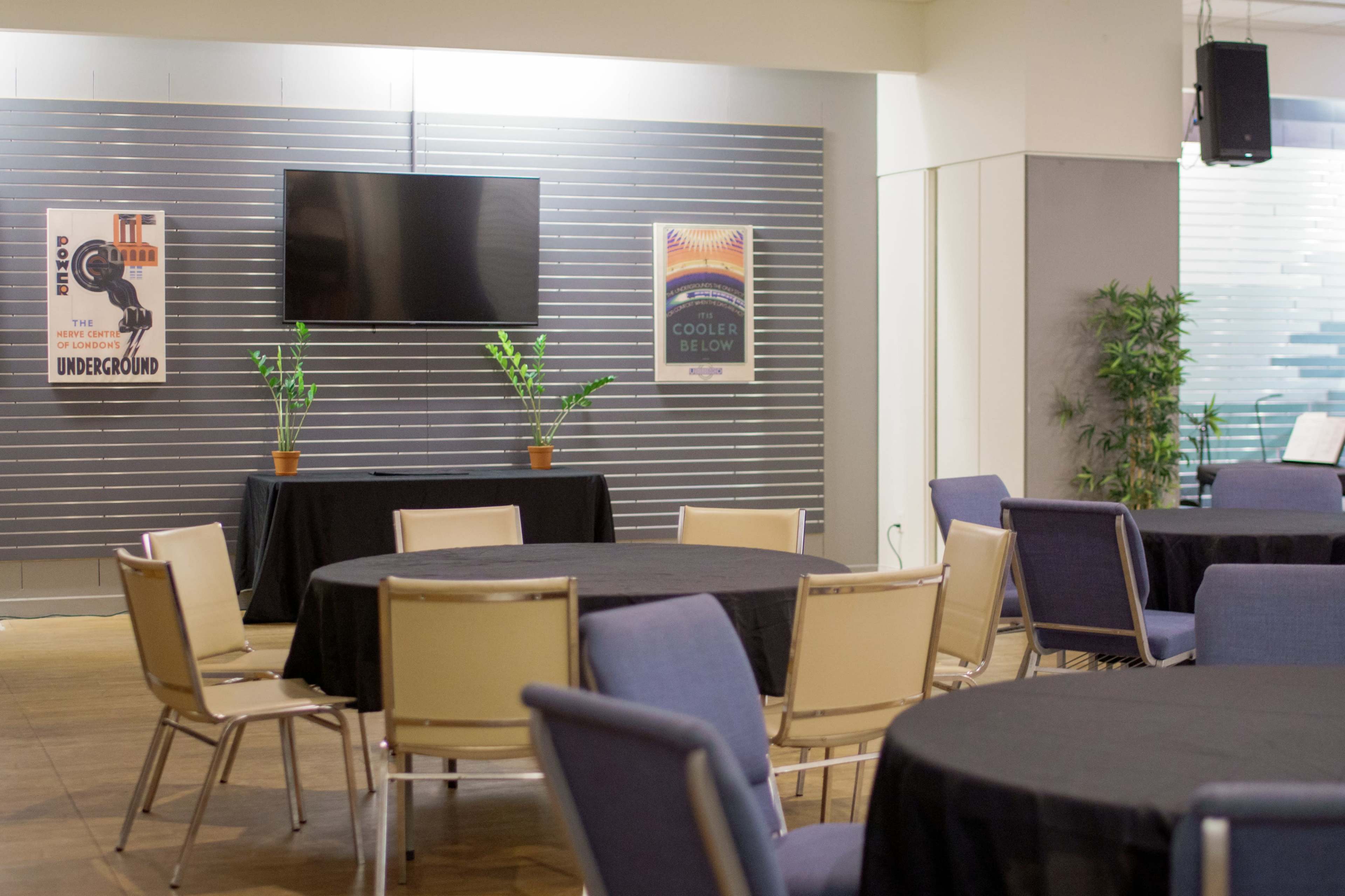 The image shows a meeting room set up with round tables covered in black cloth, a television mounted on the wall, and decorative plants in pots.