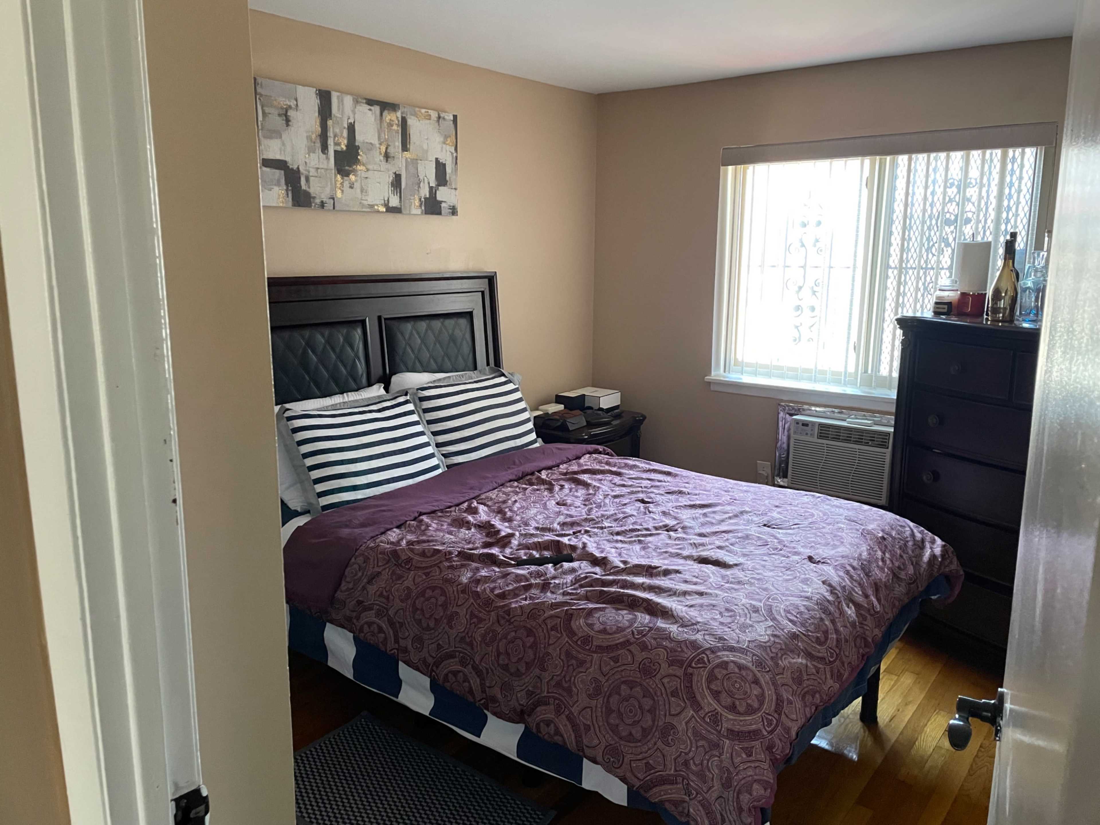 A well-lit bedroom features a bed with striped pillows and a purple comforter, accompanied by a dresser and an air conditioning unit by the window.