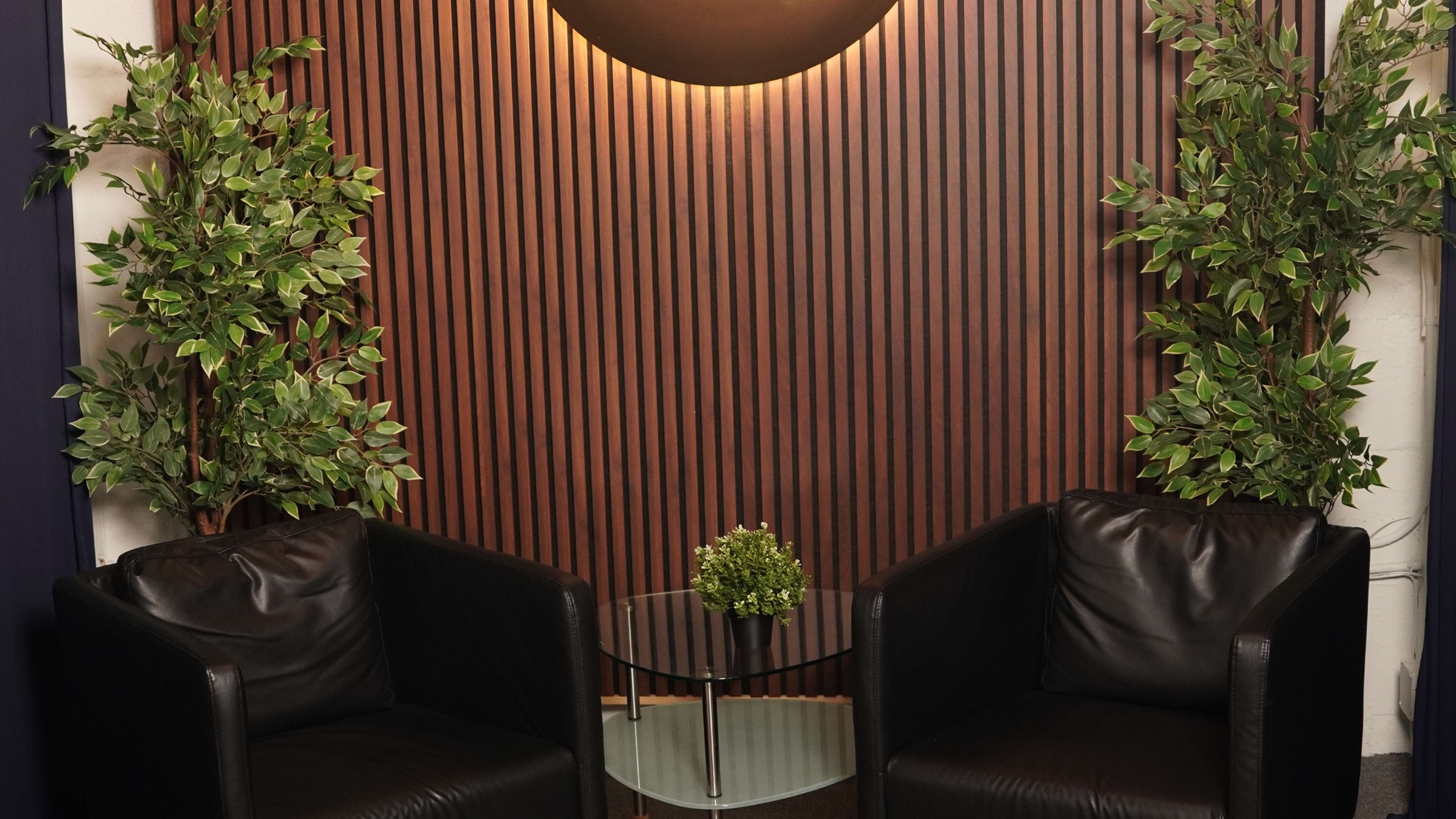 The image shows a seating area with two black leather chairs facing a small glass table, surrounded by decorative plants against a wooden slat wall.