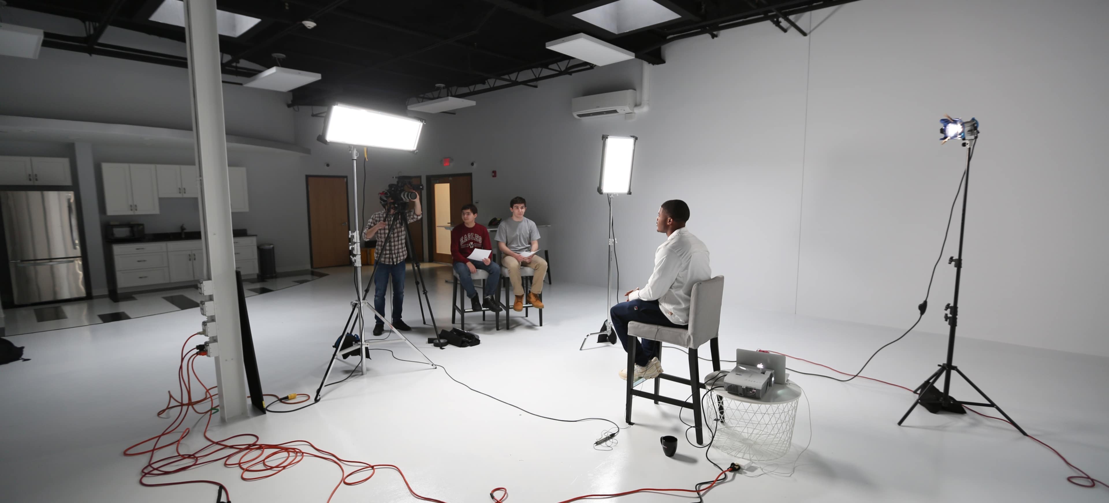 A man sits on a stool facing a camera crew in a minimalistic studio setting with bright lights and a plain background.
