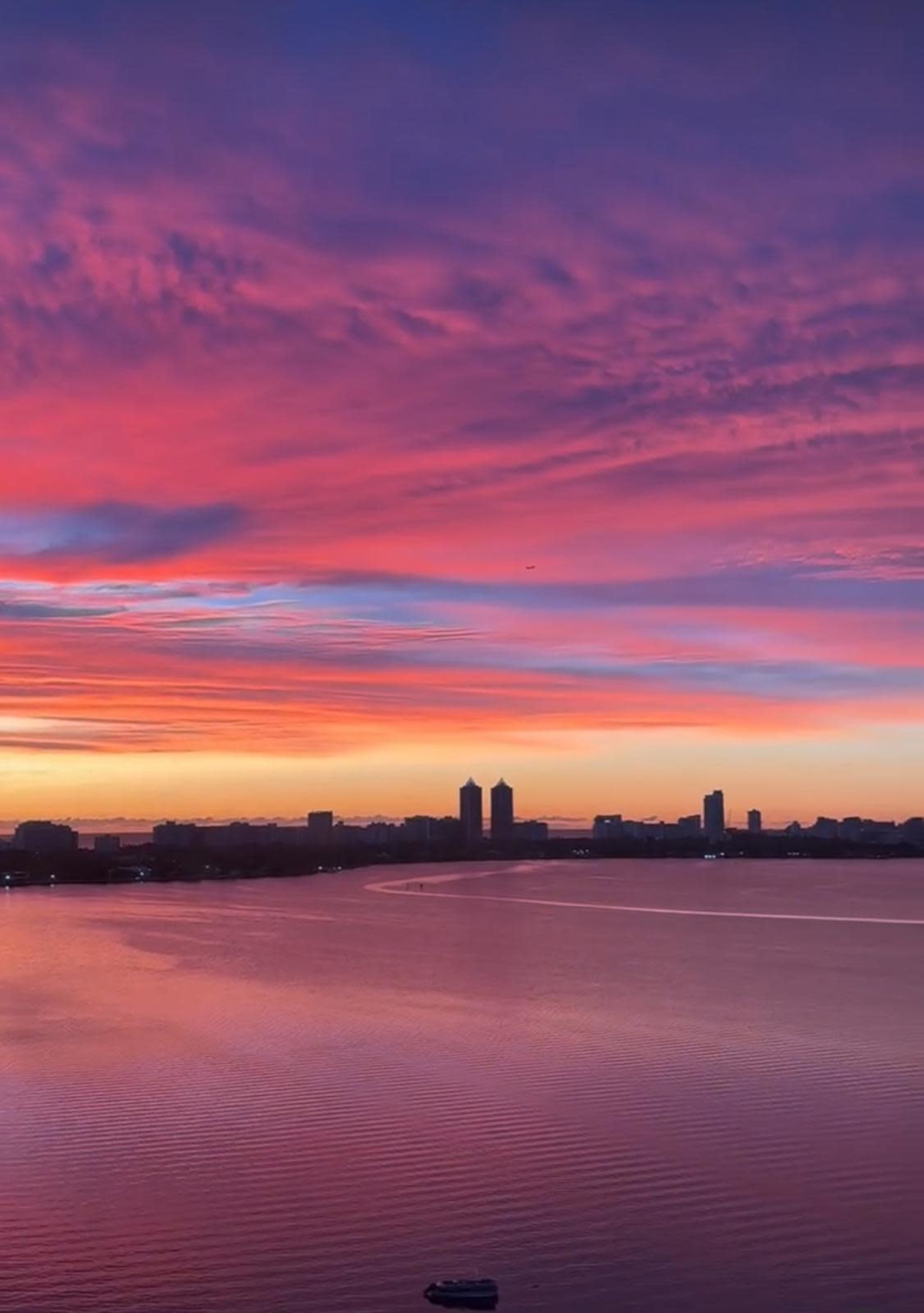 The image shows a vibrant sunset over a body of water, with silhouettes of buildings against the colorful sky.