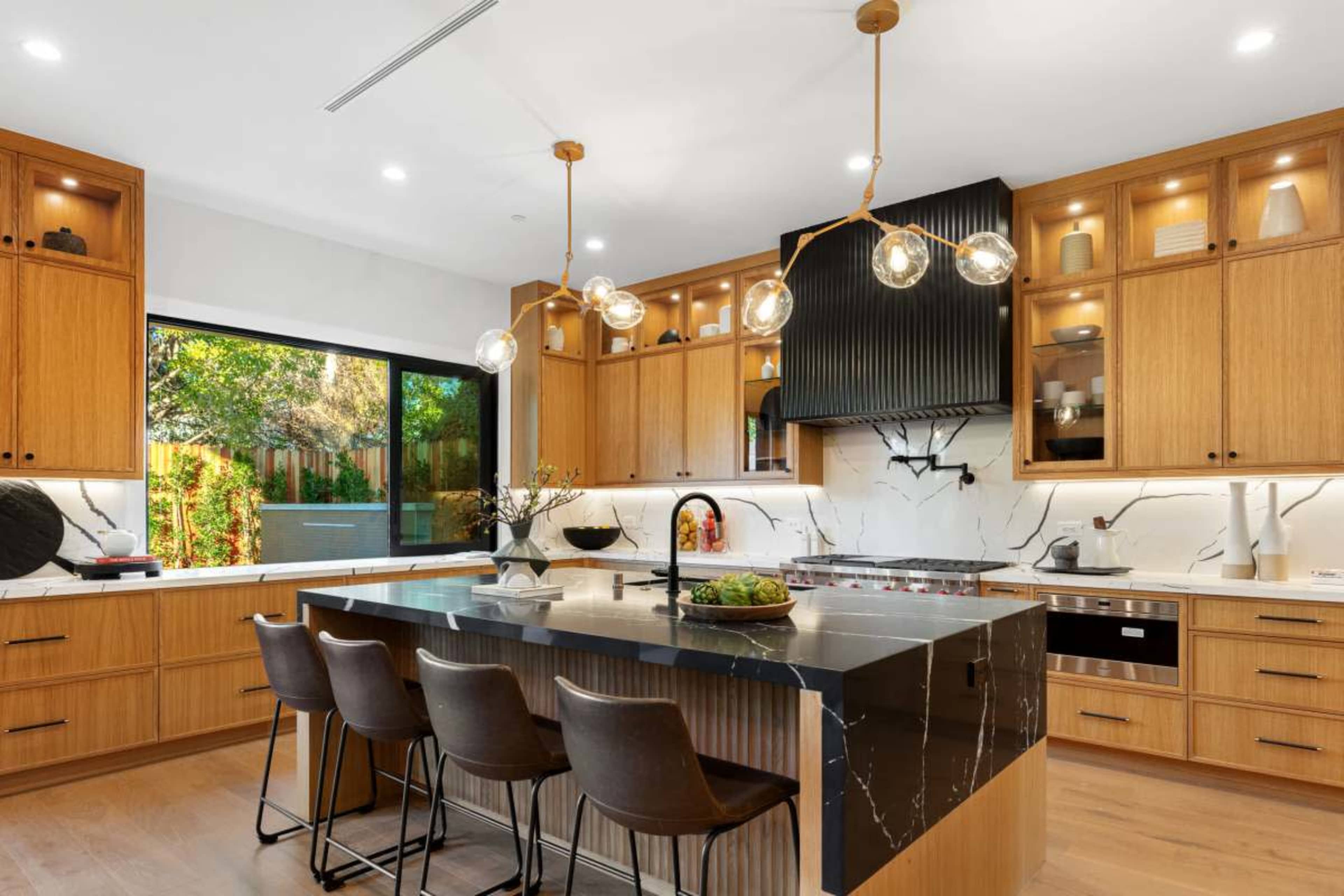 The image shows a modern kitchen with wooden cabinetry, a large island with seating, and stylish pendant lights above.