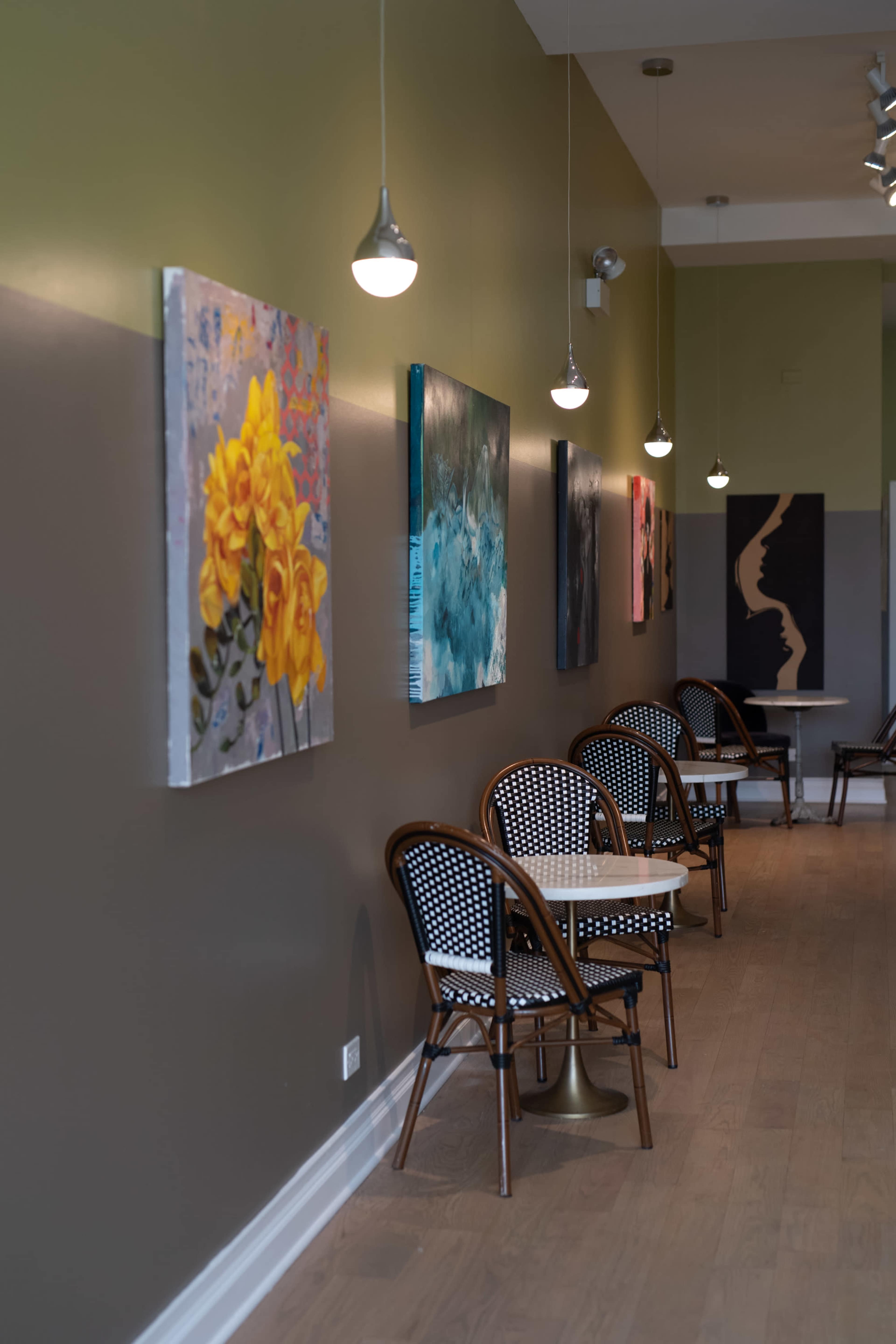 A narrow cafe interior featuring wooden chairs with patterned seats, small tables, and vibrant artwork hanging on the walls.