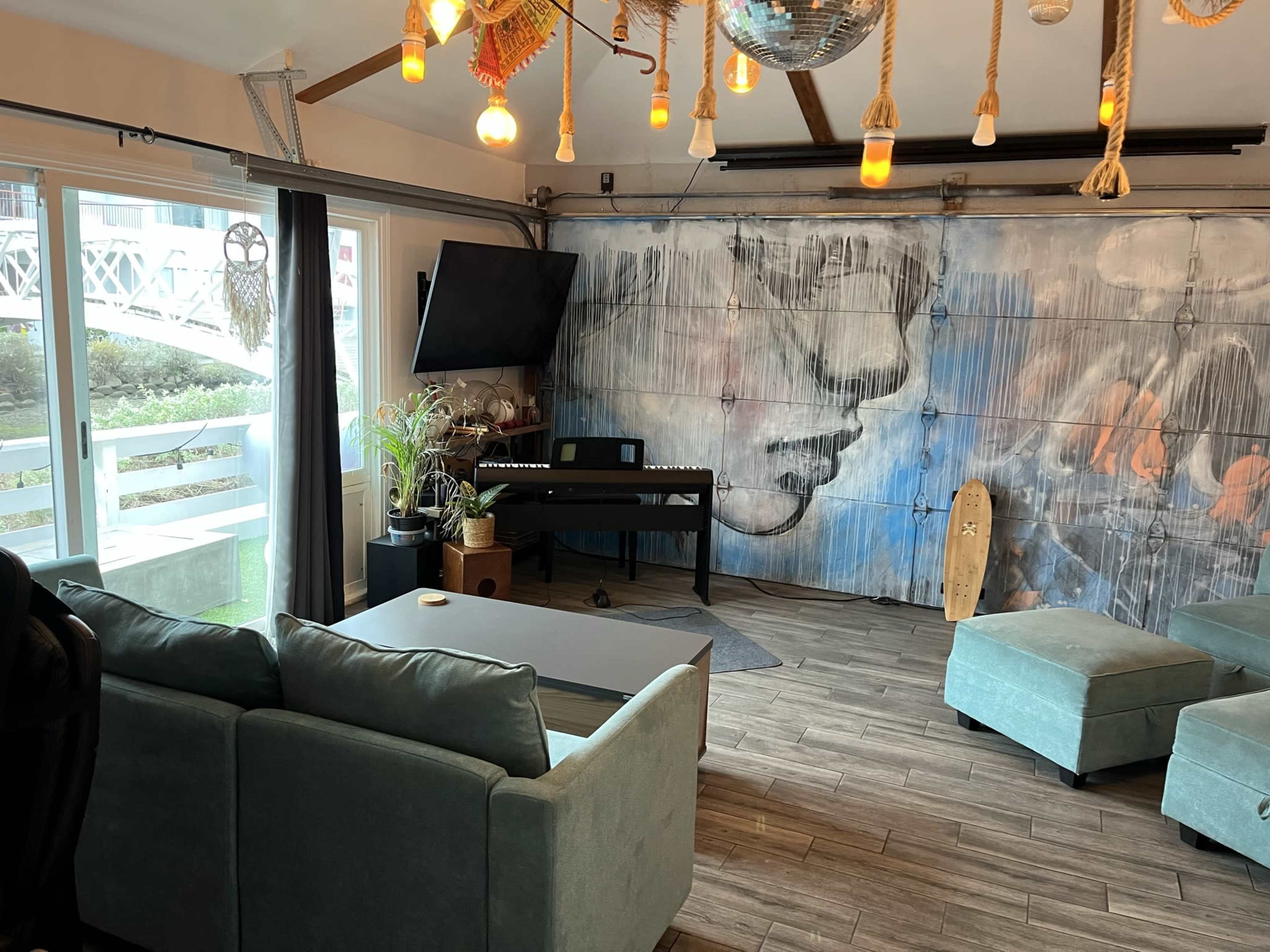 The room features a large mural of a face on one wall, a digital piano, a couch, and pendant lights hanging from the ceiling.