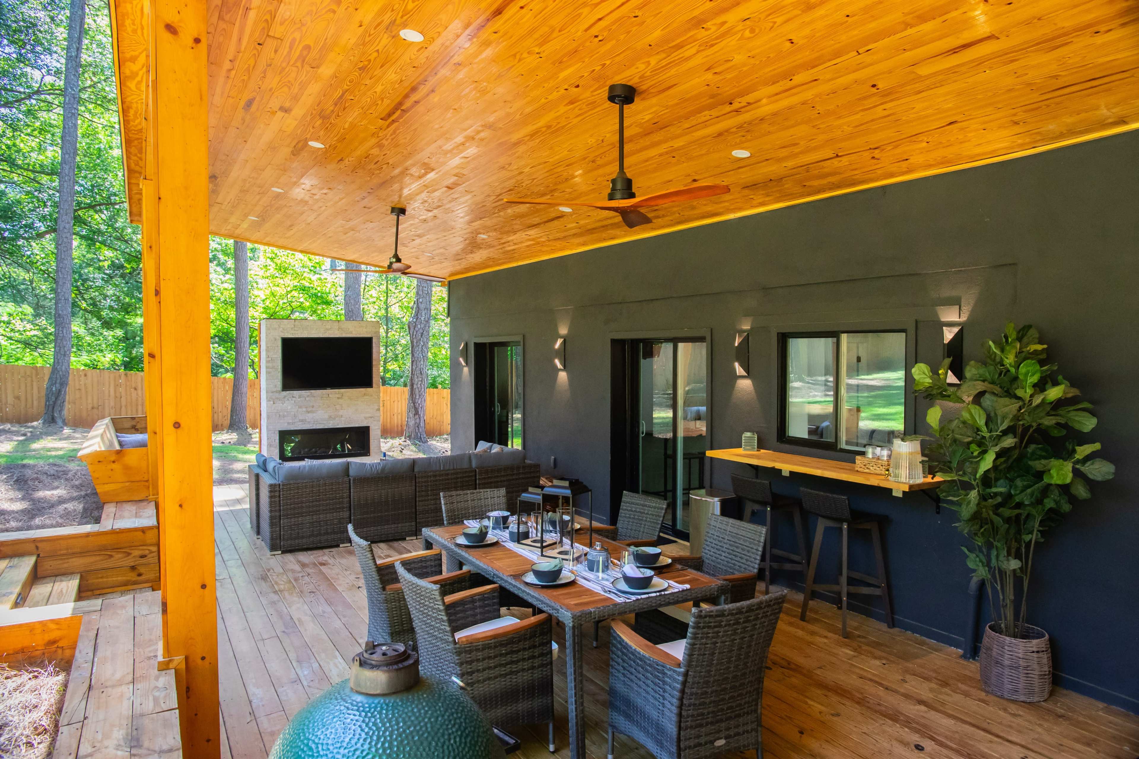 A spacious covered patio with a dining area, seating arrangement, and a large outdoor television, surrounded by trees.