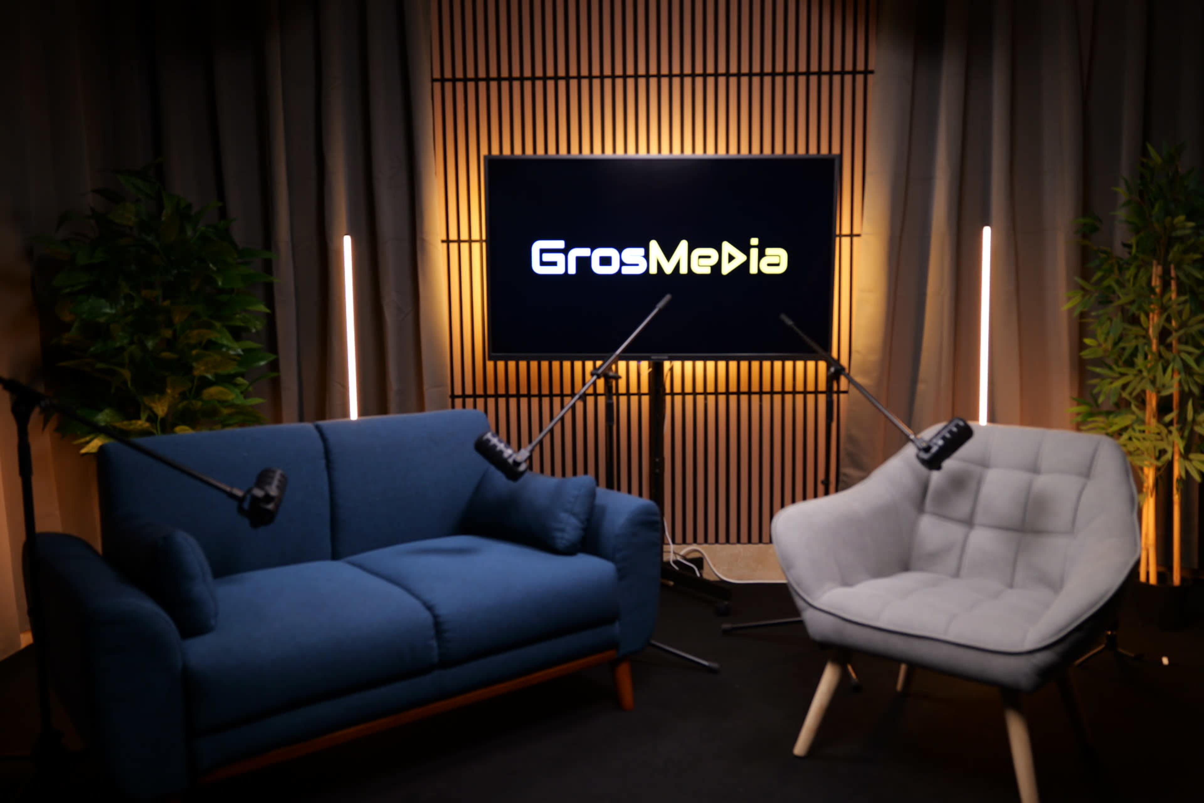 The image shows a modern studio setup featuring a blue sofa and a gray chair, both positioned in front of a large screen displaying the logo "GrosMedia," with studio lights and plants in the background.