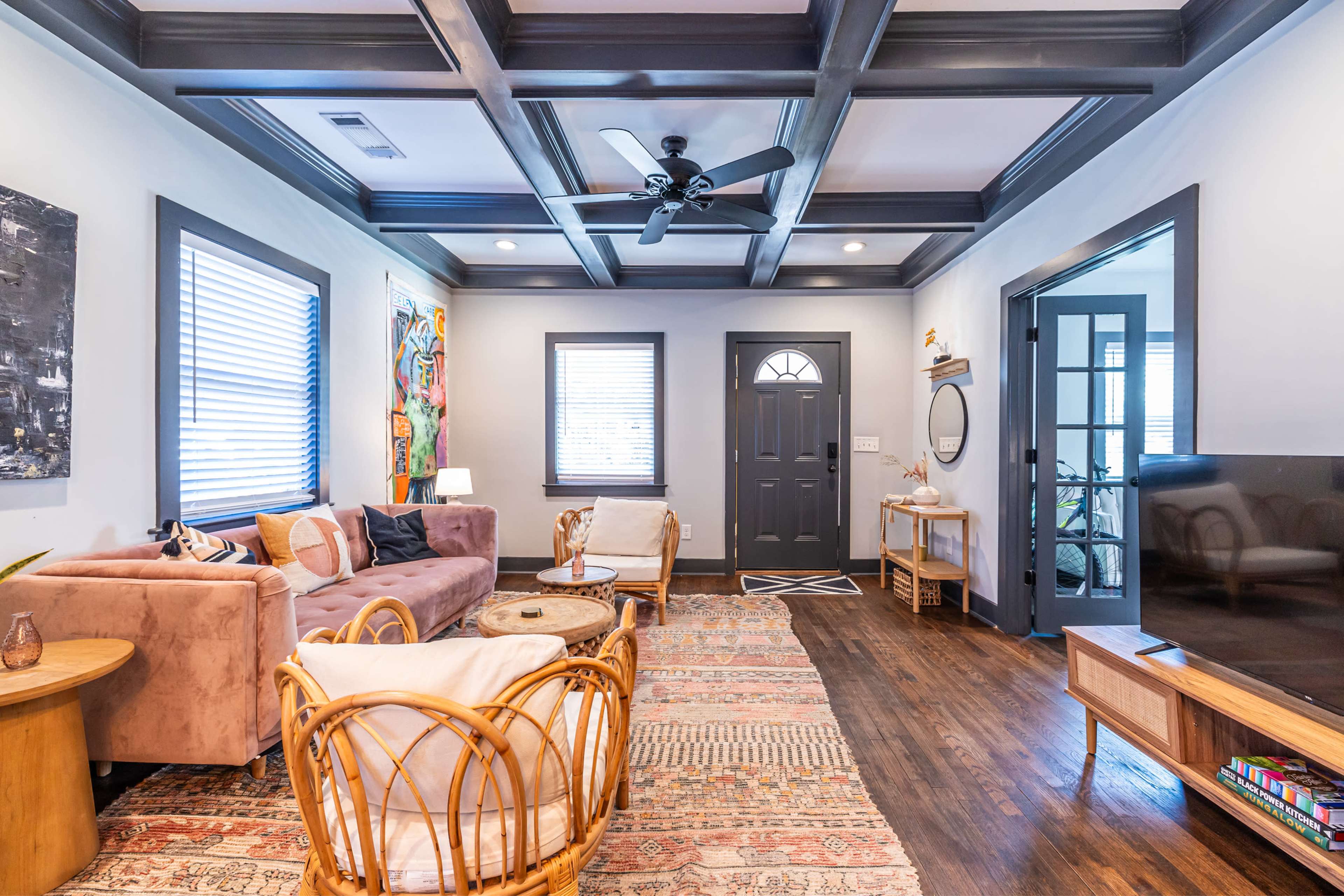 The living room features a beige sofa, a patterned rug, a coffee table, and a dark front door, with a mix of wall art and large windows.
