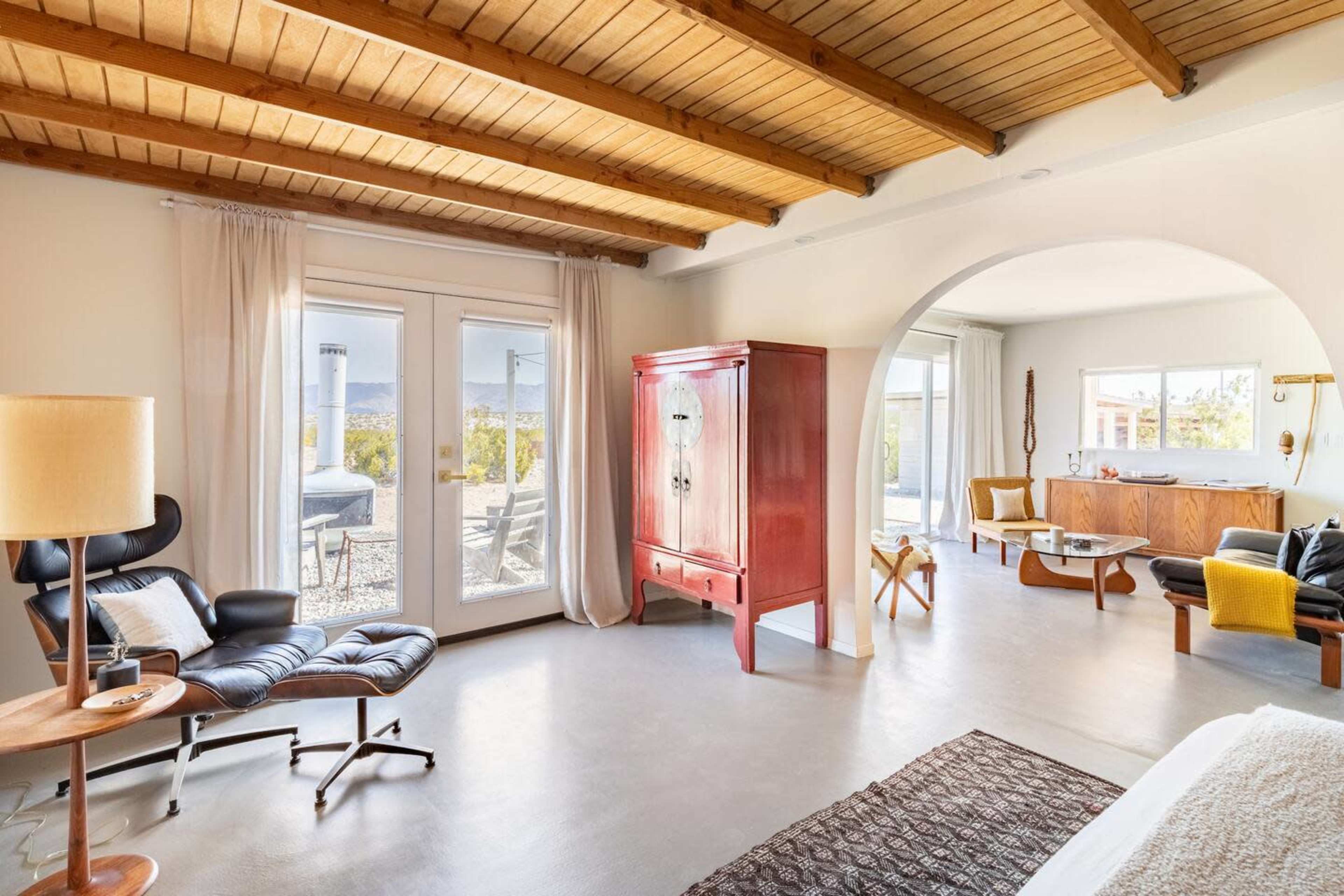 The image features a modern living room with wooden beams on the ceiling, a red cabinet, and large windows offering a view of the outdoors.