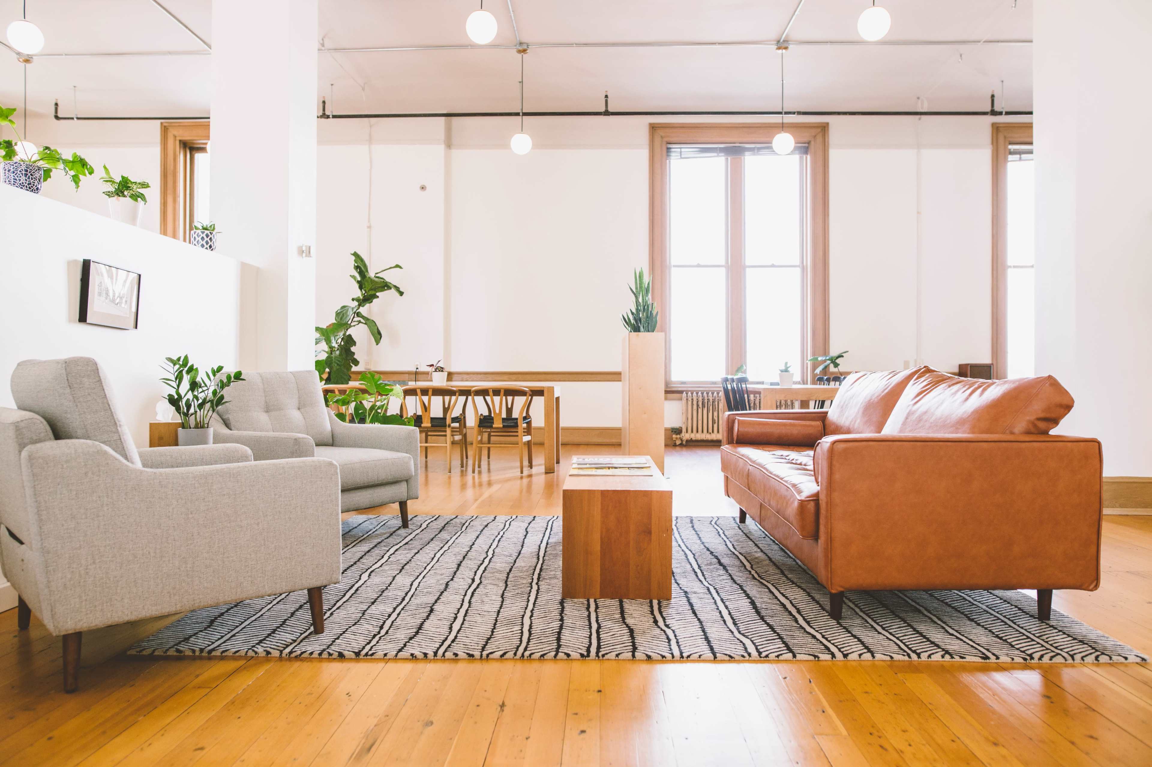A bright, modern living area features a brown leather sofa, two light gray armchairs, a wooden coffee table, and a dining area with several chairs against a backdrop of indoor plants.