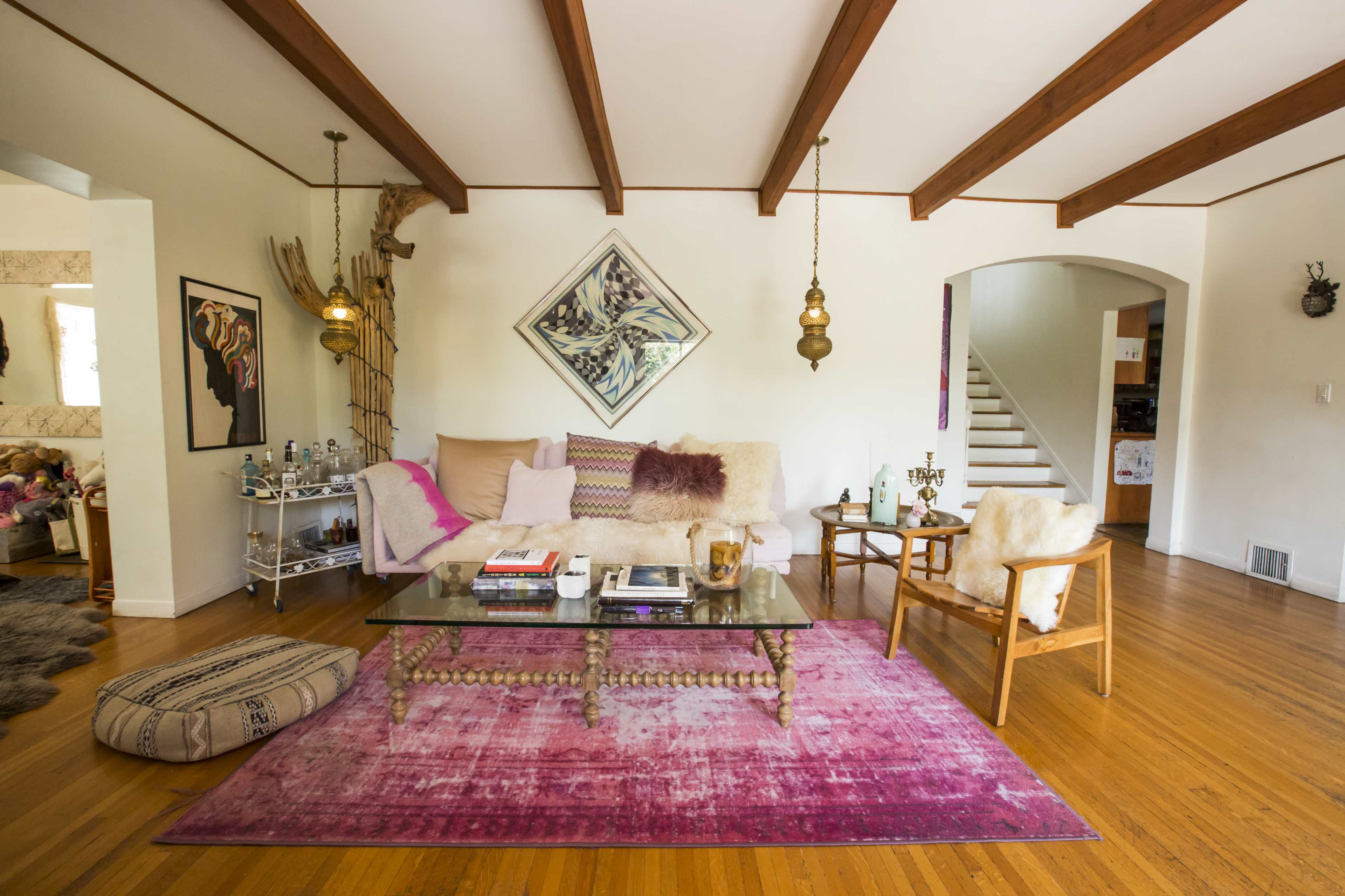 A modern living room featuring a pink area rug, wooden beams on the ceiling, a glass coffee table, and stylish seating arrangements.
