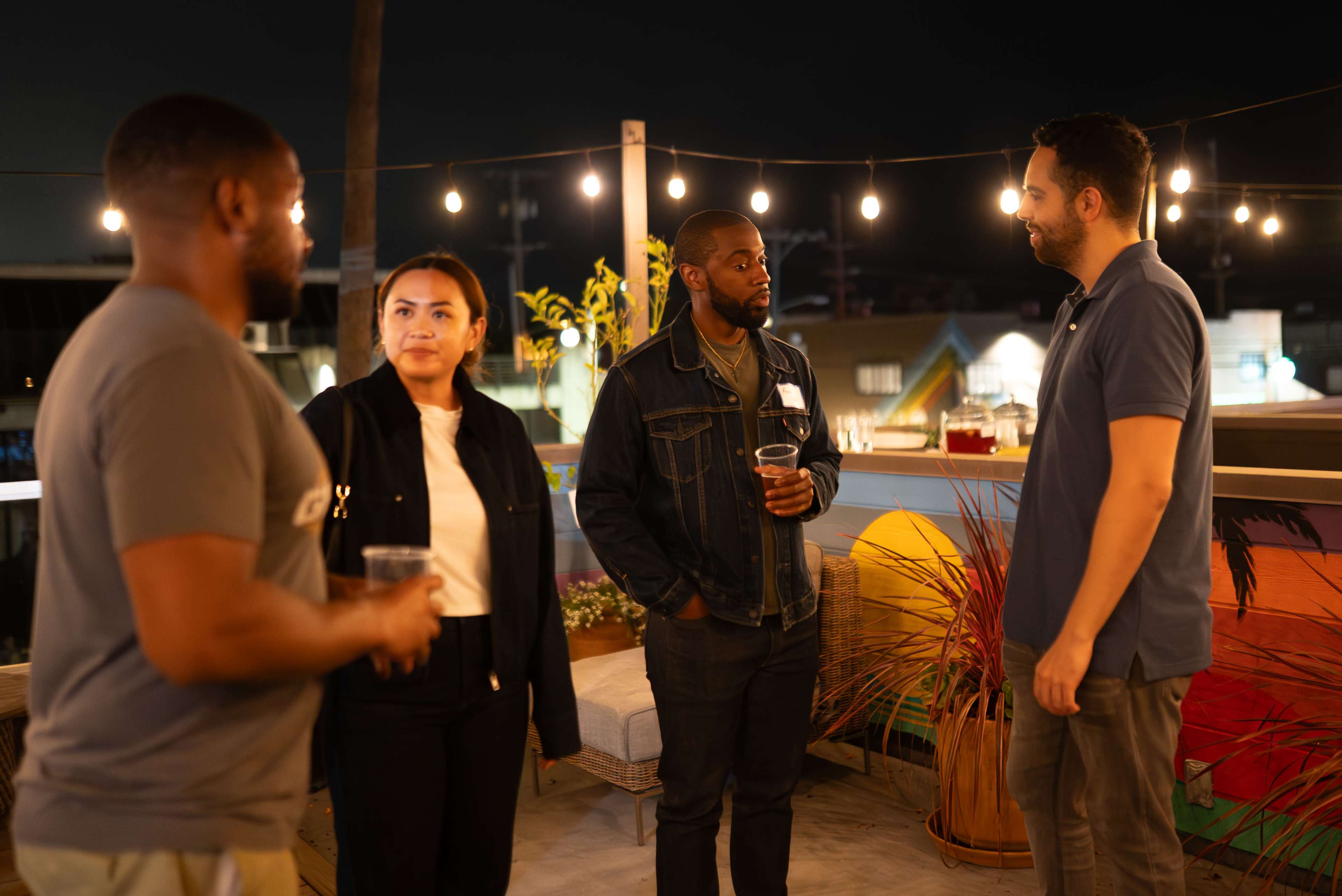 A group of four people is having a conversation on a rooftop at night, surrounded by string lights and potted plants.