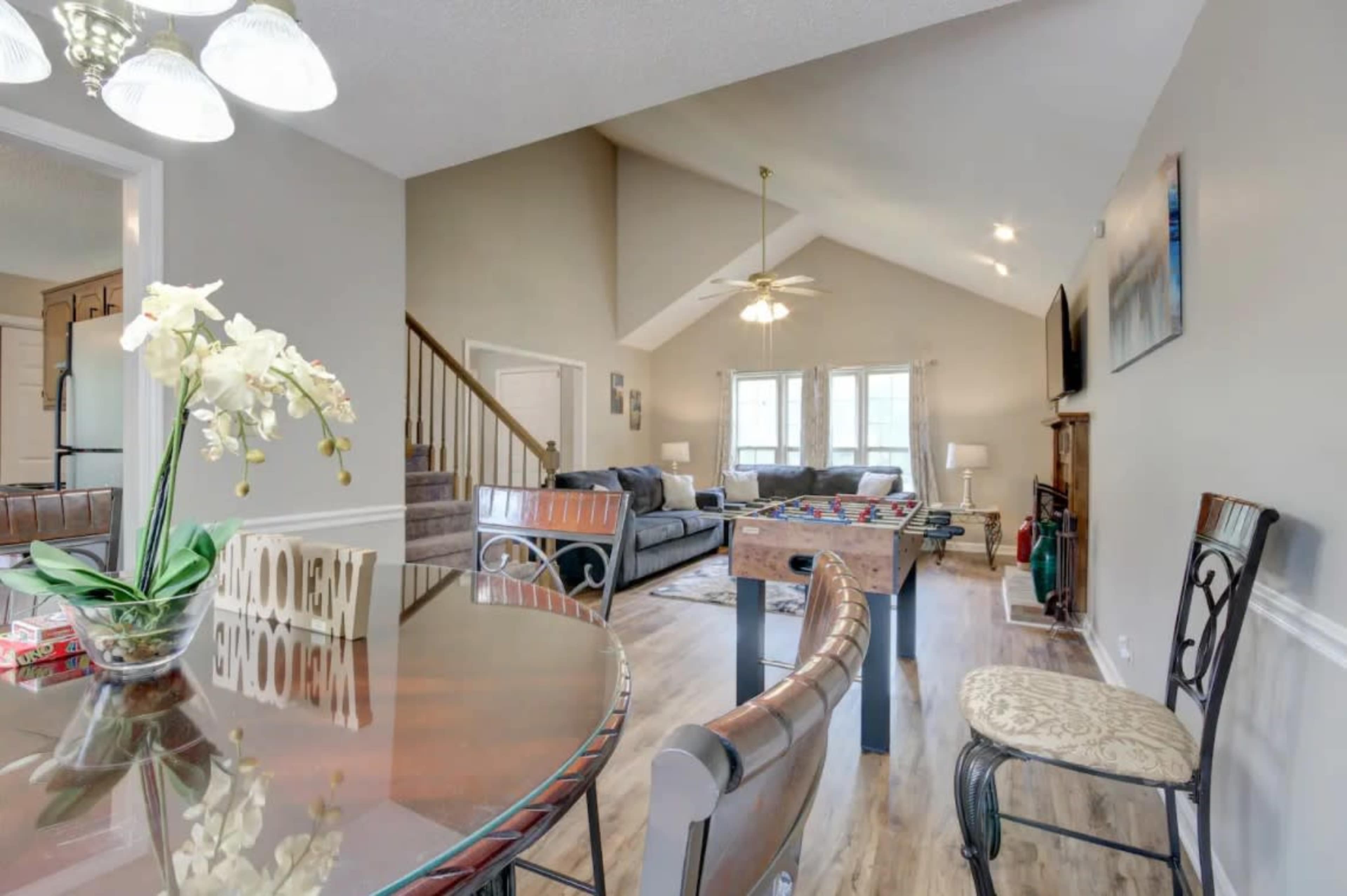 The image shows a spacious living area with a dining table in the foreground, a foosball table in the middle, and a comfortable seating area with a large sofa in the background.