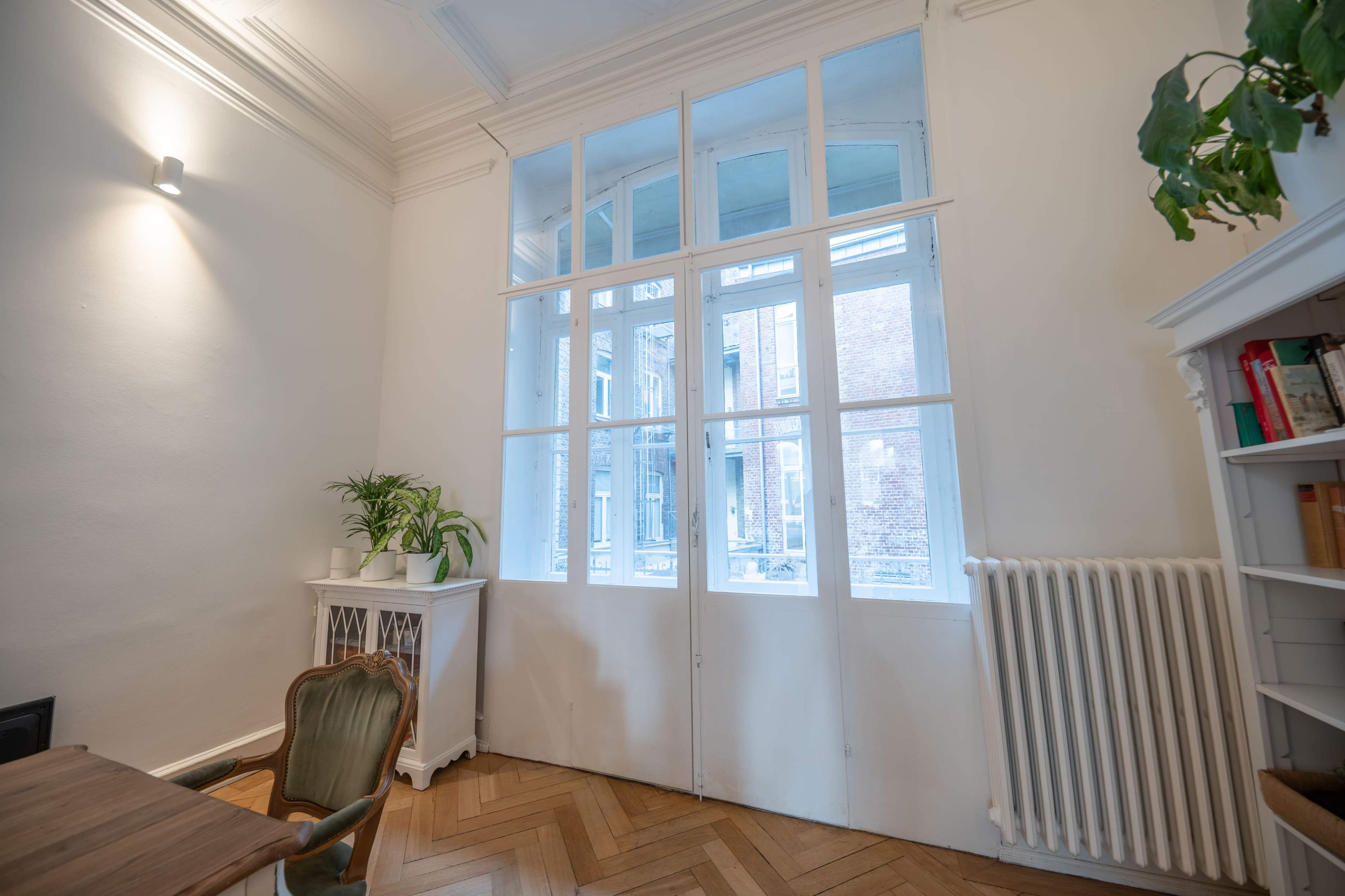 The image shows a bright room with large windows, a wooden table, a chair, and a white bookshelf, featuring a potted plant and a radiator along the wall.