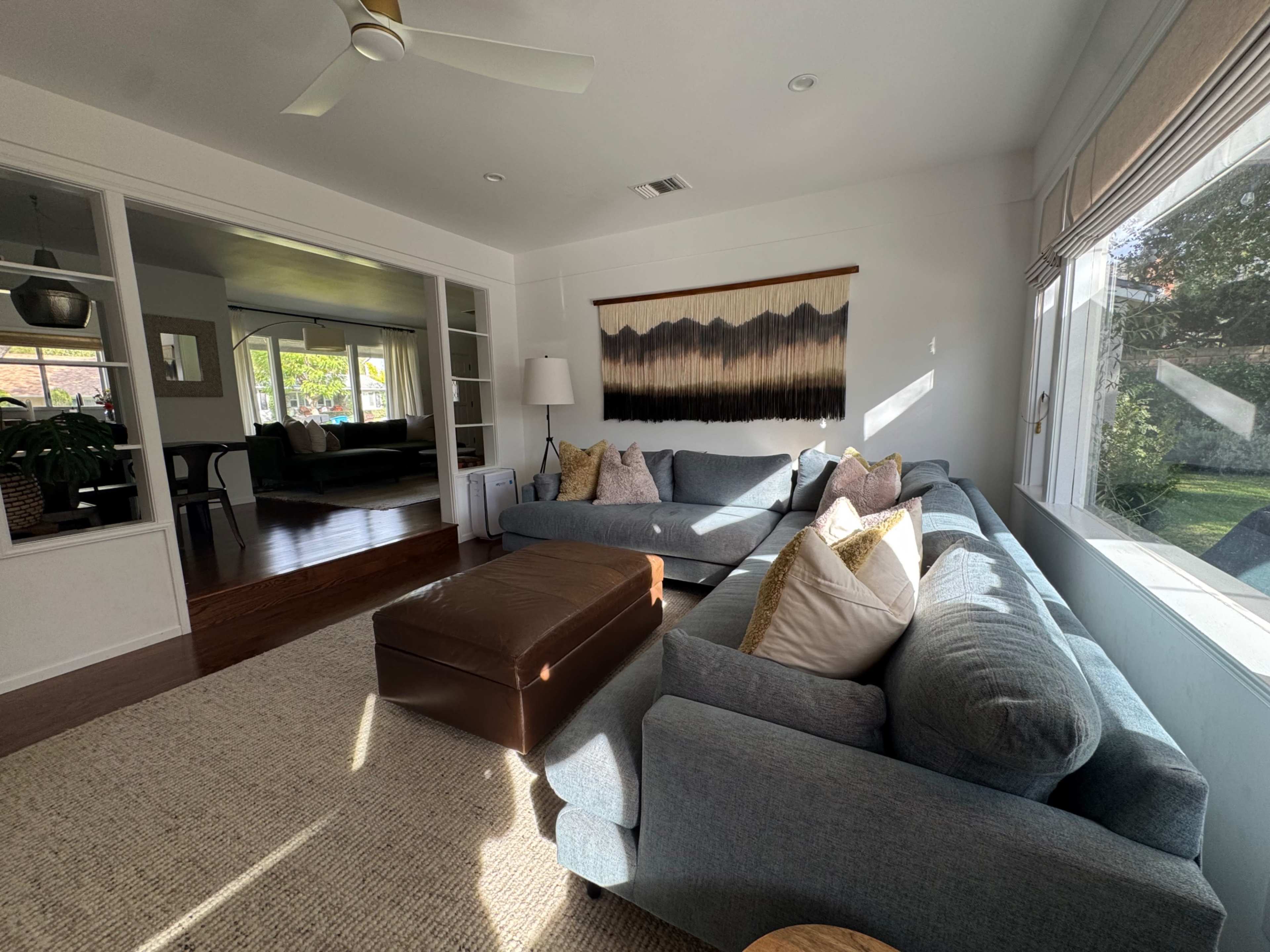 A cozy living room features a large gray sectional sofa adorned with pillows, a brown ottoman, and a wall hanging, all with abundant natural light streaming through the windows.