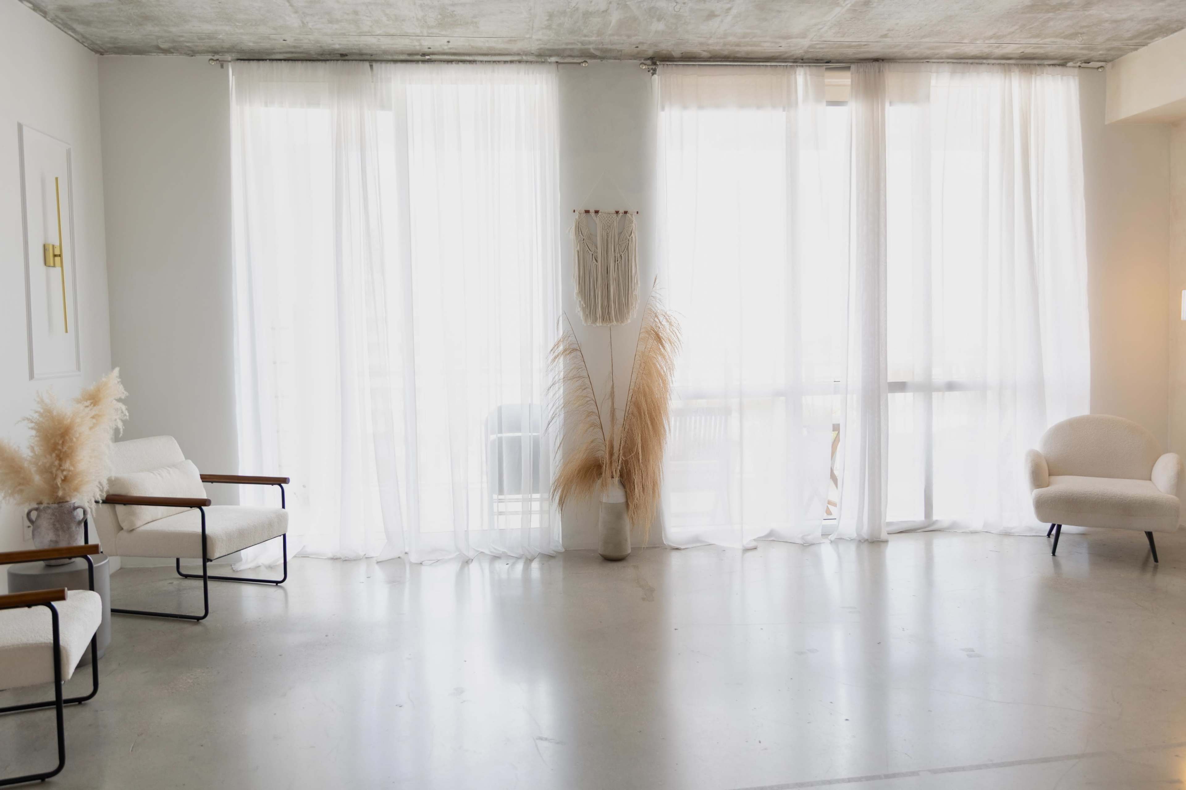 The image features a minimalistic interior space with large sheer curtains, two chairs, and a decorative plant in a vase.