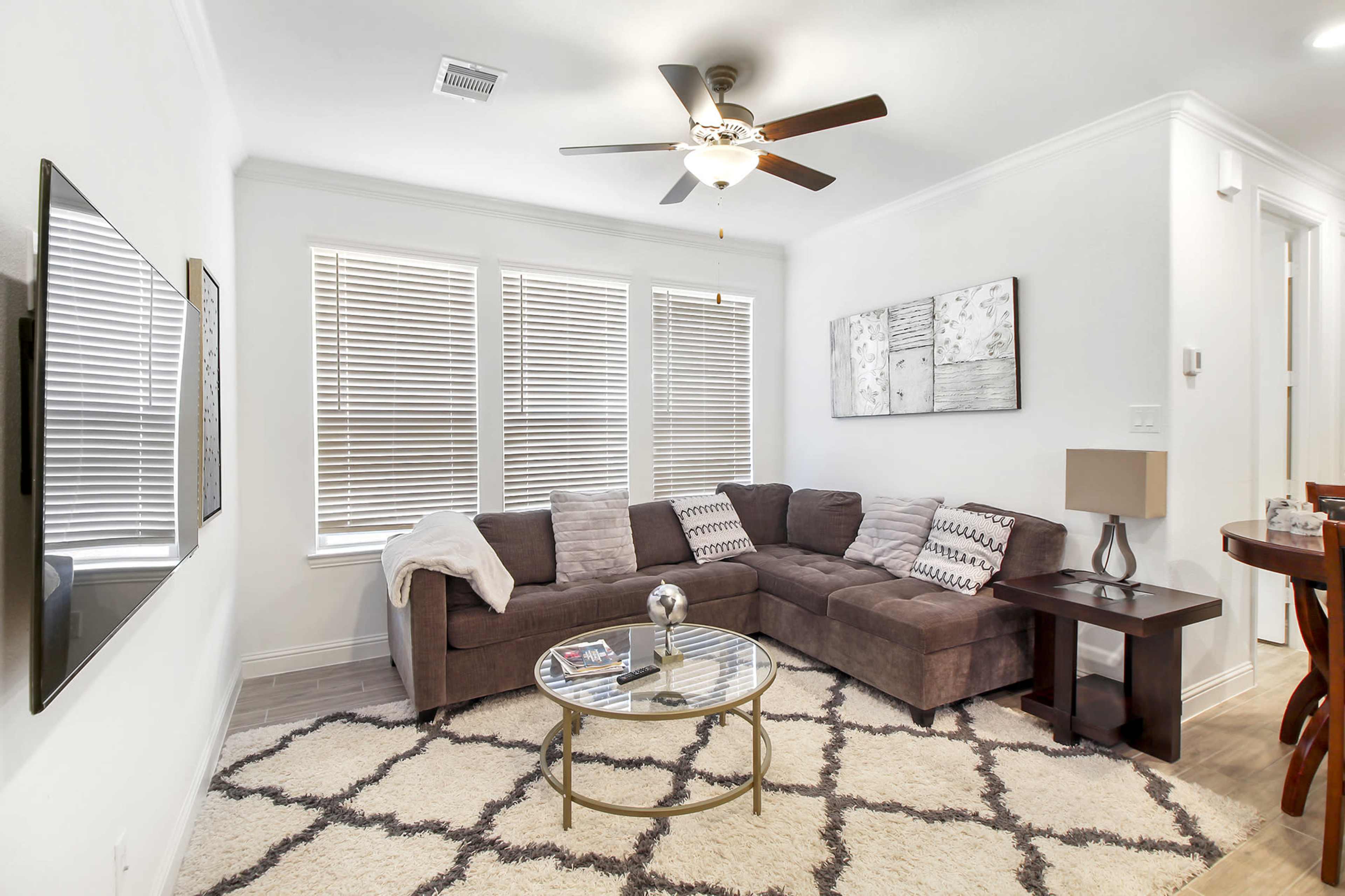 A modern living room features a sectional sofa, a glass coffee table, and a television mounted on the wall, with sunlight streaming through the window blinds.
