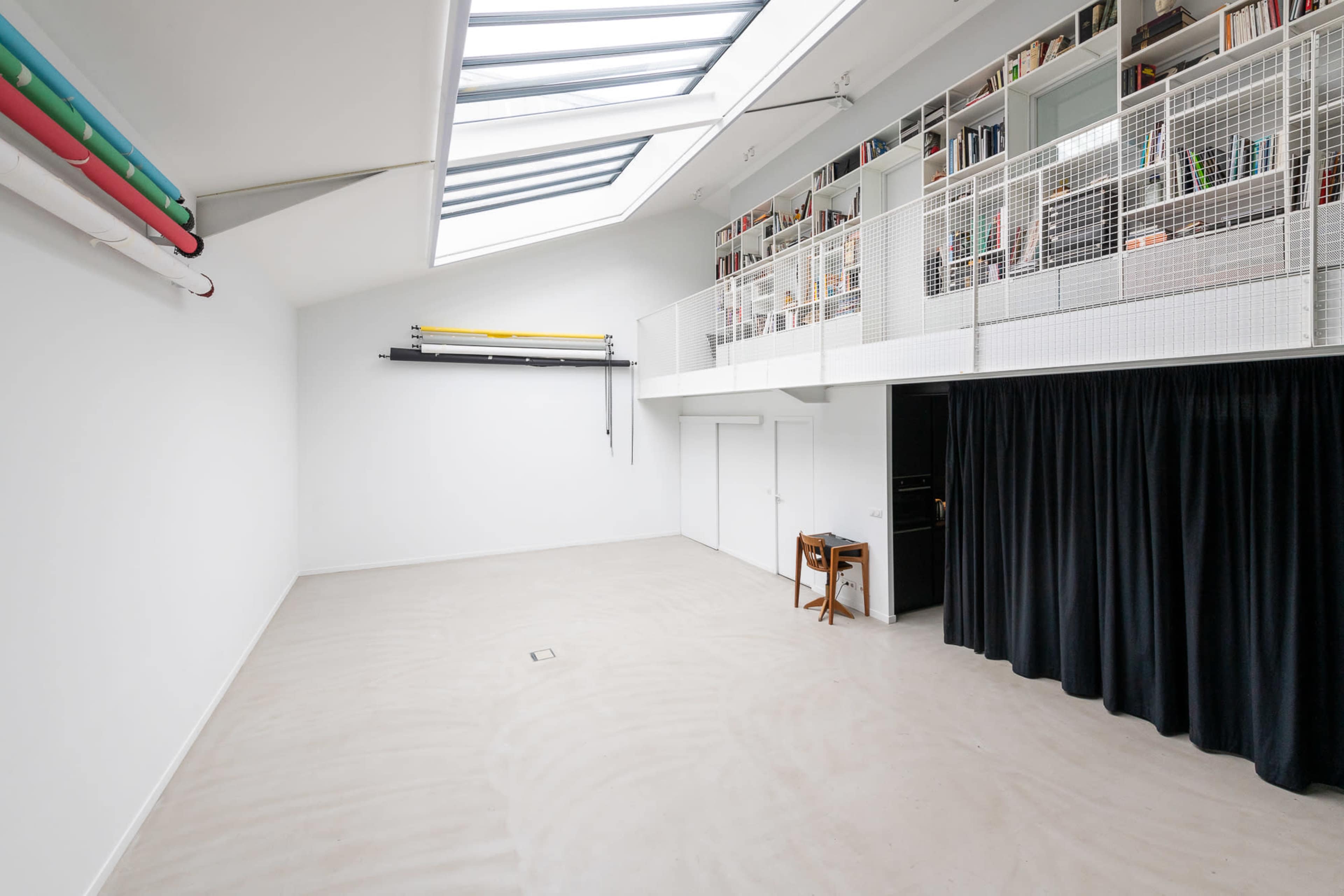 An empty room with a skylight, white walls, a storage loft with books, and a black curtain on one side.