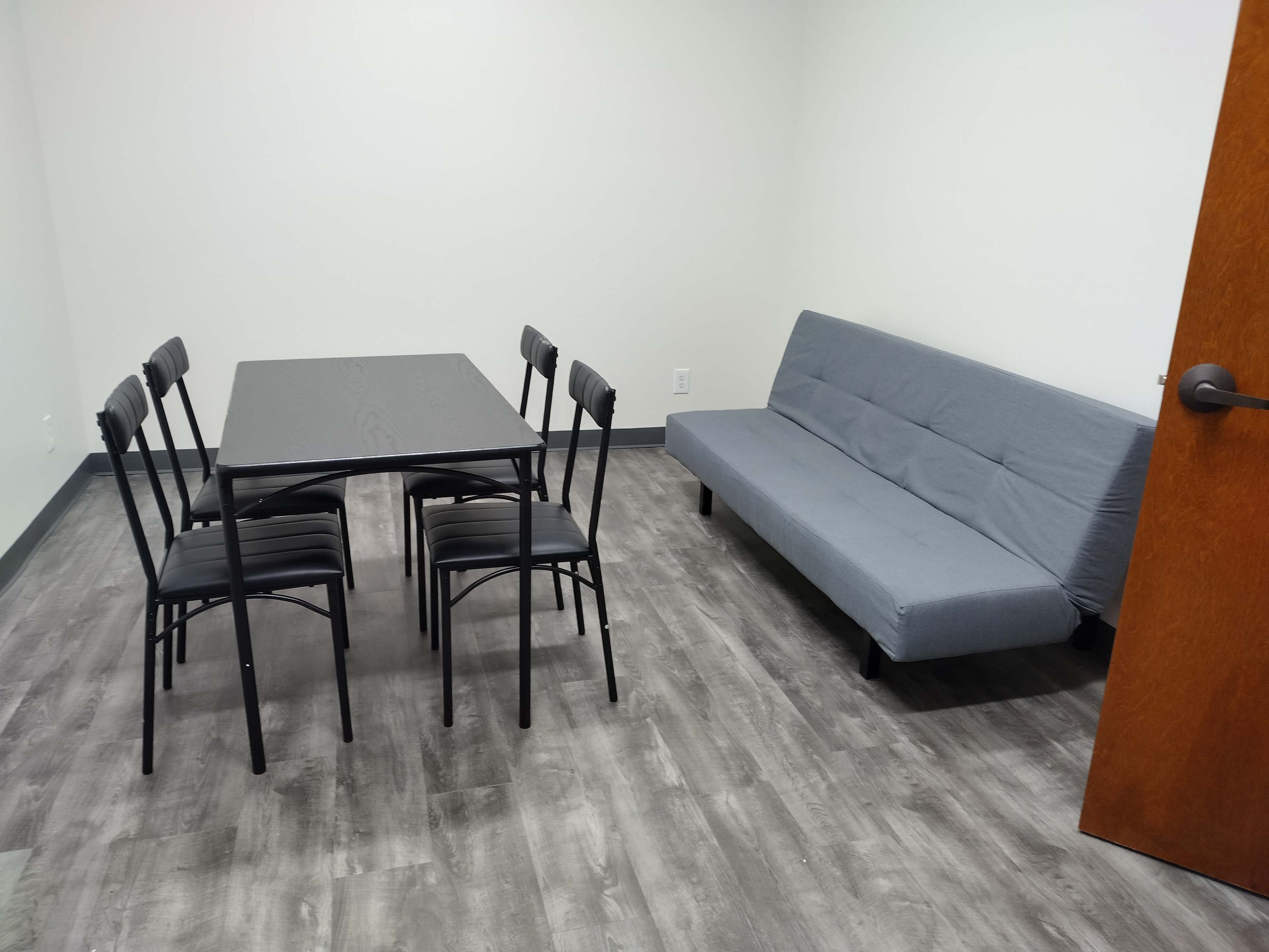 The image shows a small, empty room containing a gray futon on one side and a black dining table with six chairs on the other.