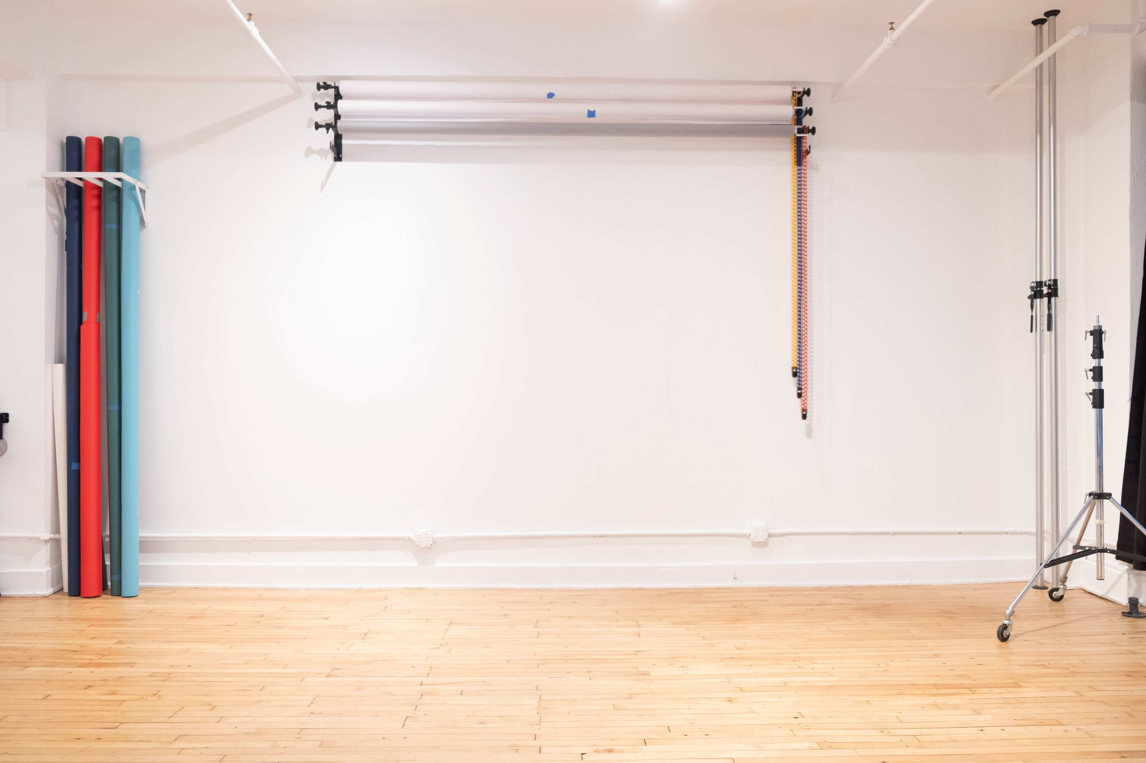 The image shows a minimalist studio space with various backdrop rolls mounted on the wall and colored poles leaning against one side.