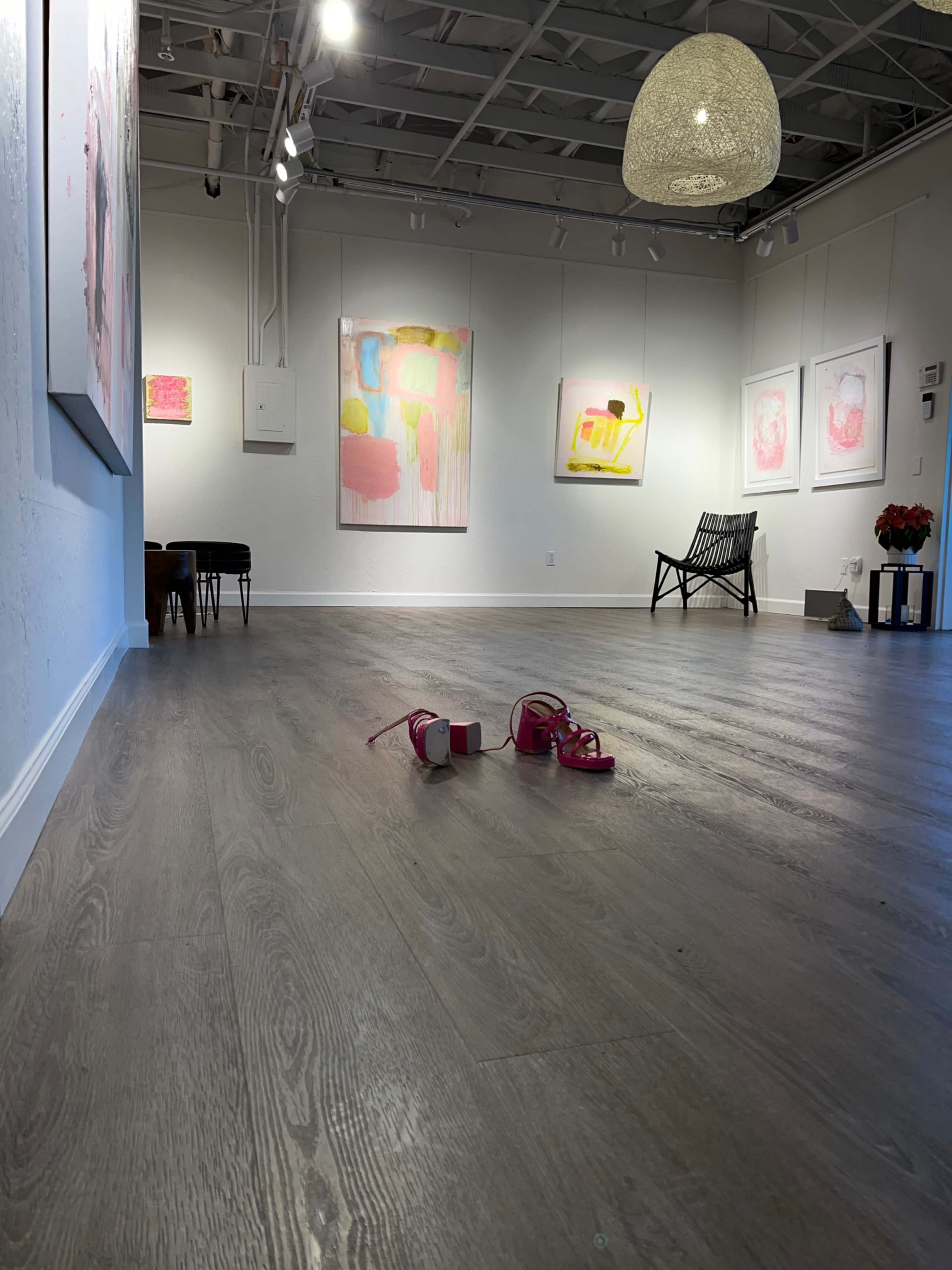 A spacious art gallery with colorful abstract paintings on the walls, a flower arrangement on the floor, and a pair of pink sandals off to the side.
