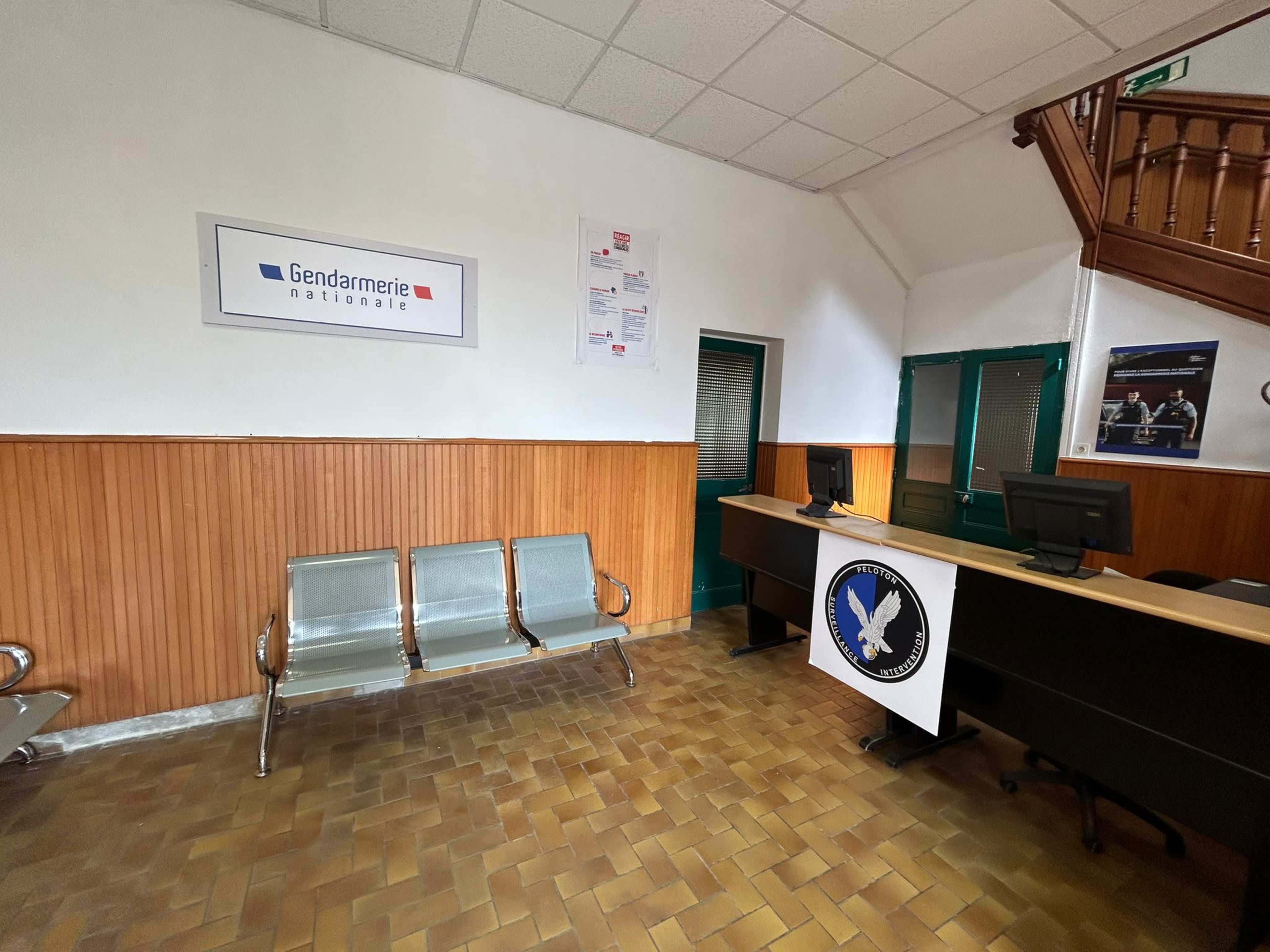 The interior of a gendarmerie office, featuring a reception desk with computers and metal benches against a wooden wall.