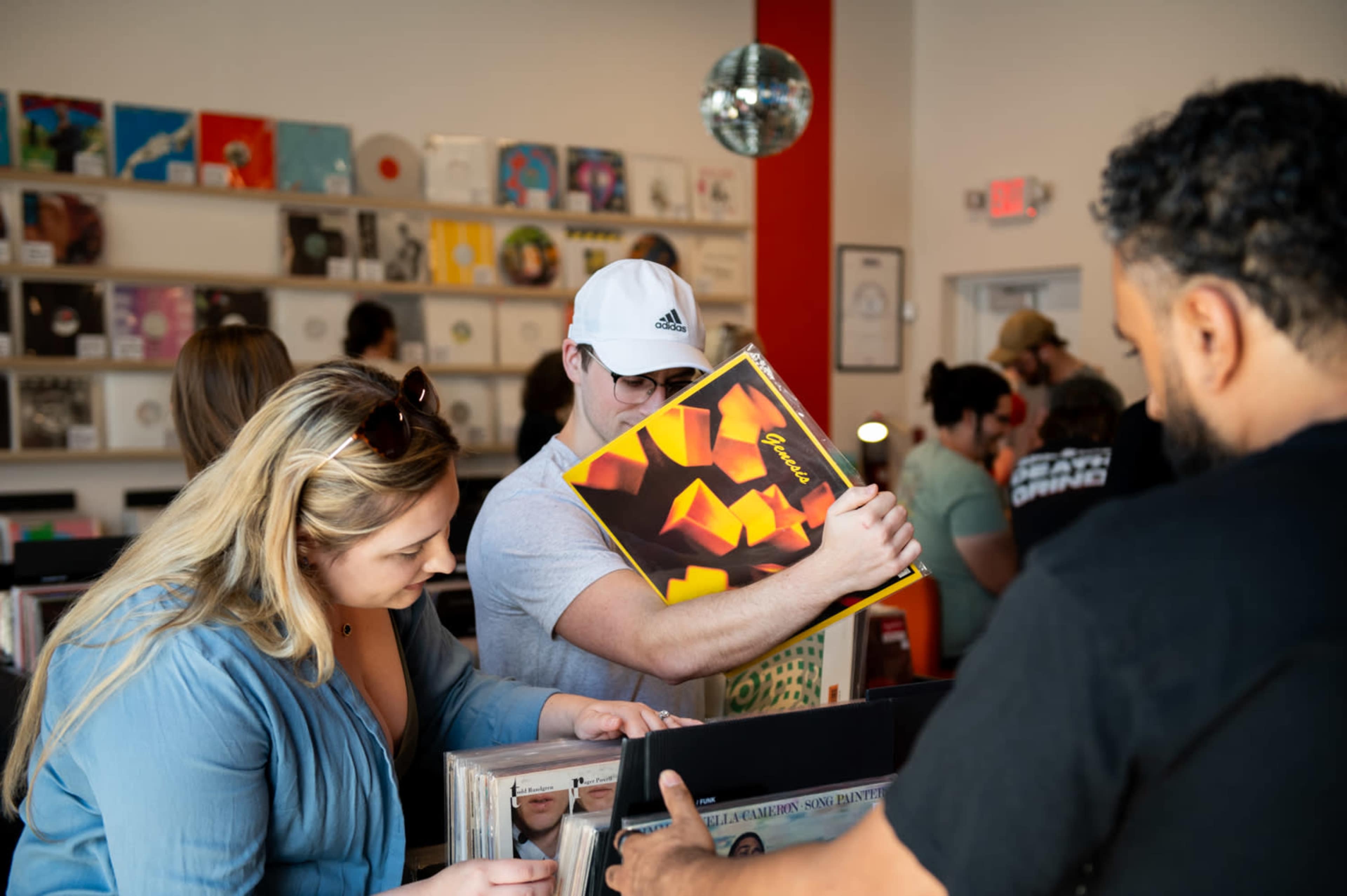 Two people browse through a selection of vinyl records in a record store, while others look on in the background.
