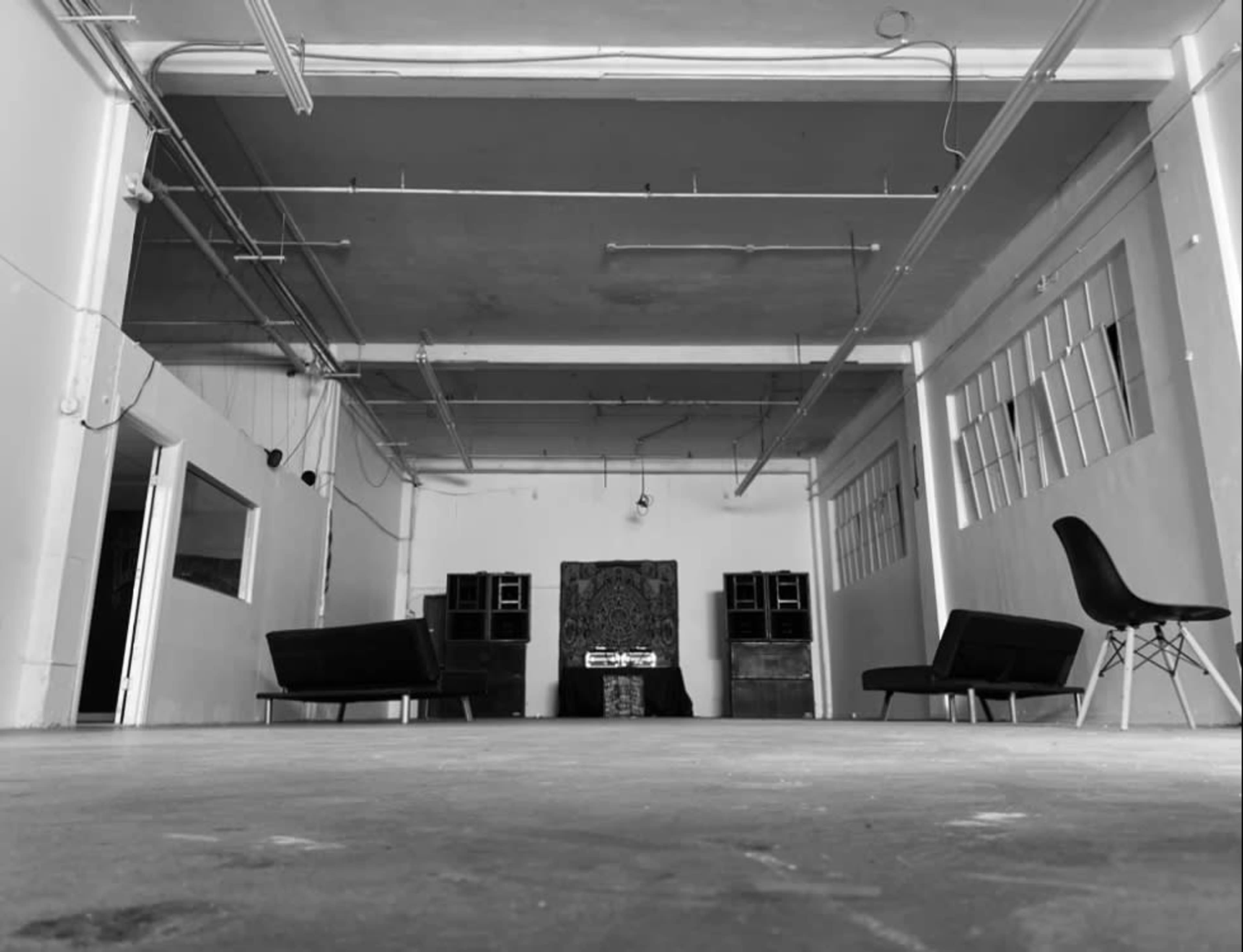 The image shows a spacious, empty room with two black sofas facing a central area that features a decorative piece and sound equipment.