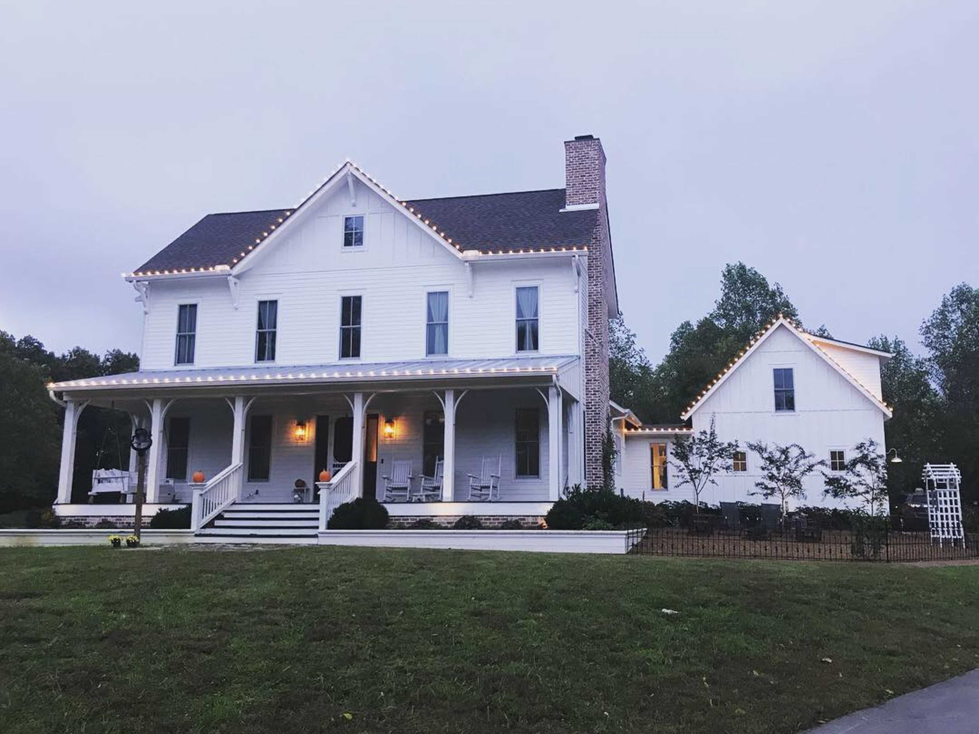 A two-story white house with a gabled roof features string lights along the eaves and a porch surrounded by a landscaped yard.