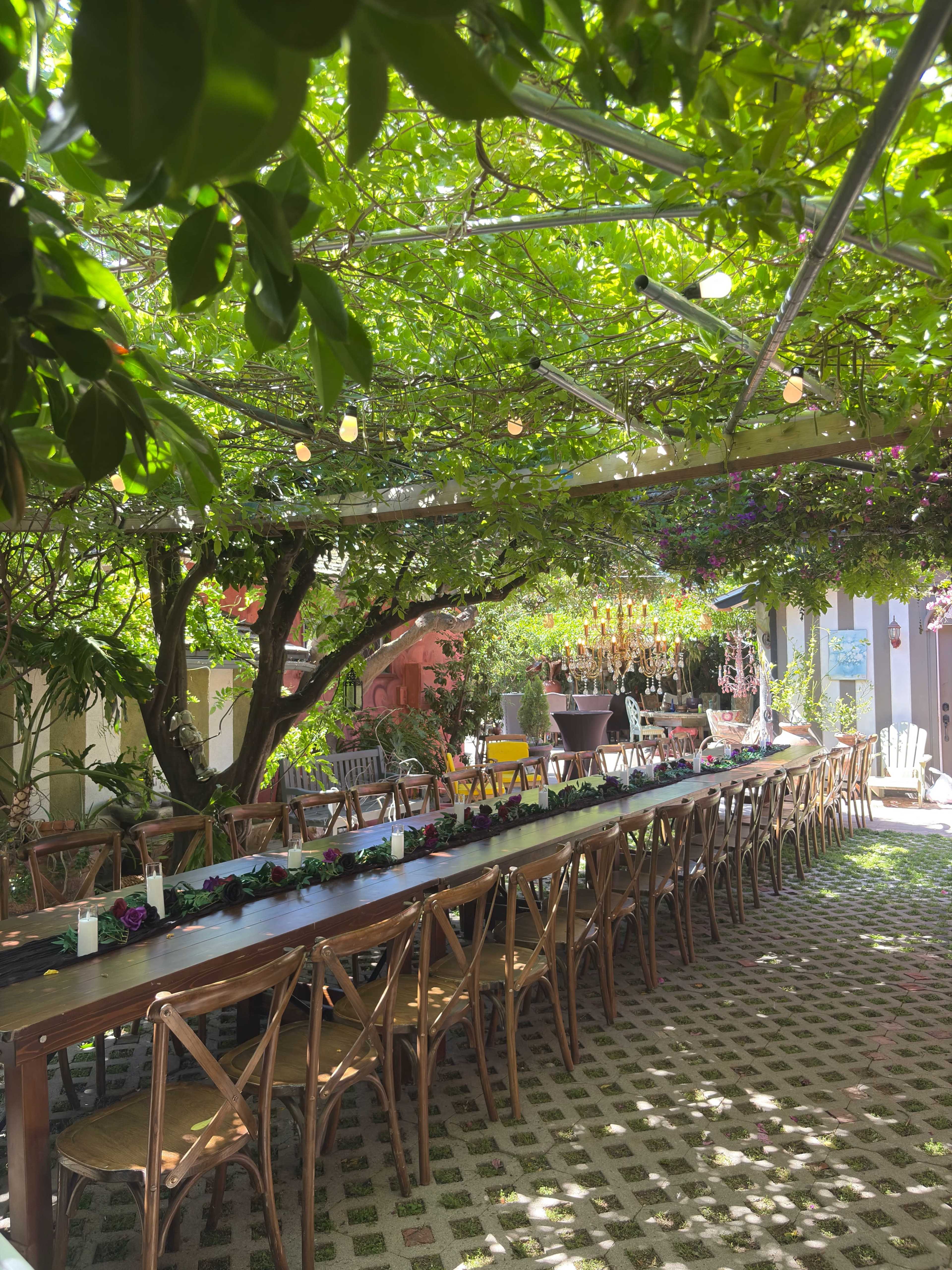 A long wooden table is set under a canopy of lush green vines, surrounded by chairs and decorated with floral arrangements in a shaded outdoor space.