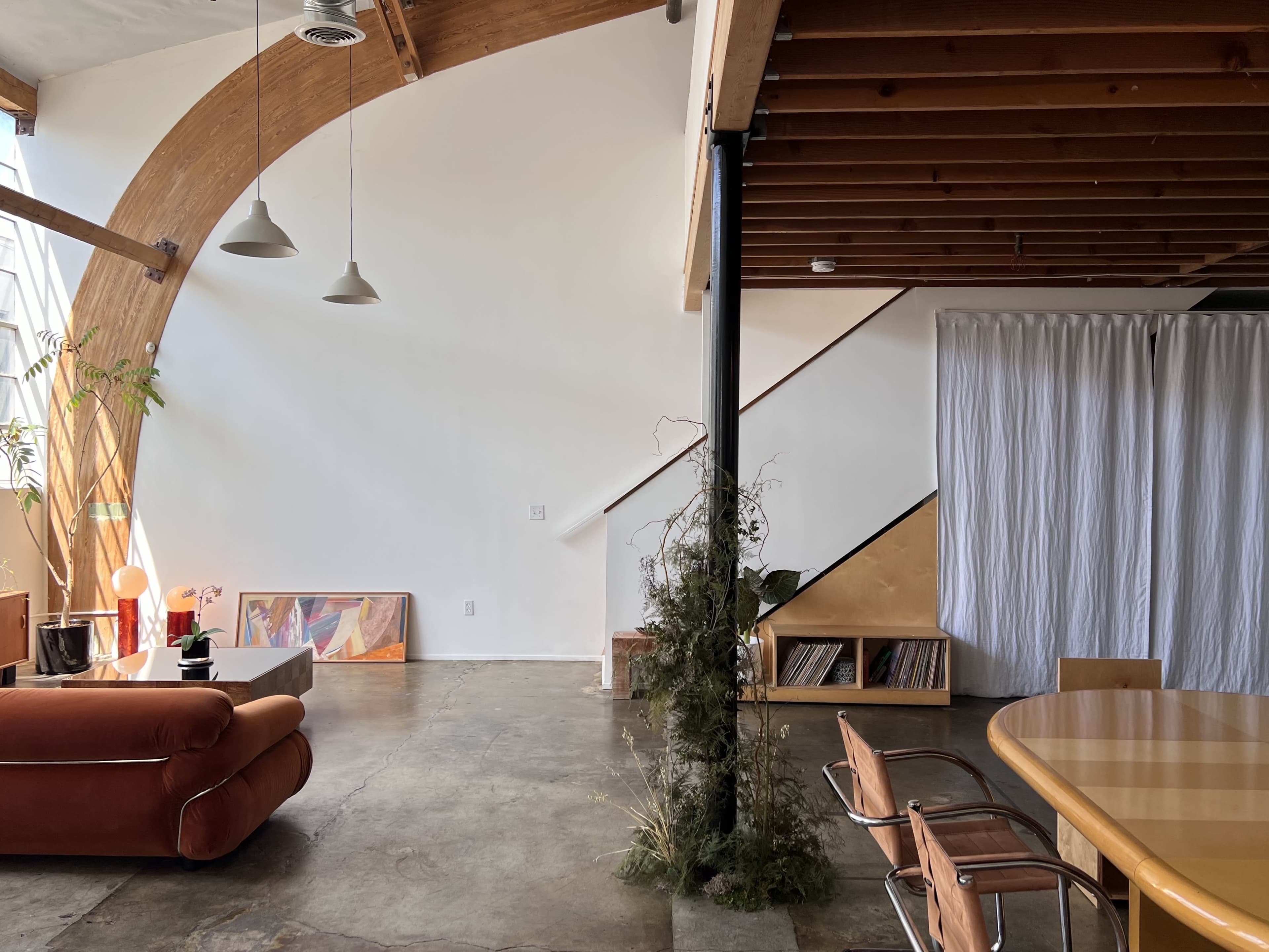 An open interior space featuring a curved wooden beam, a sofa, a dining table, and a set of stairs with a curtain.