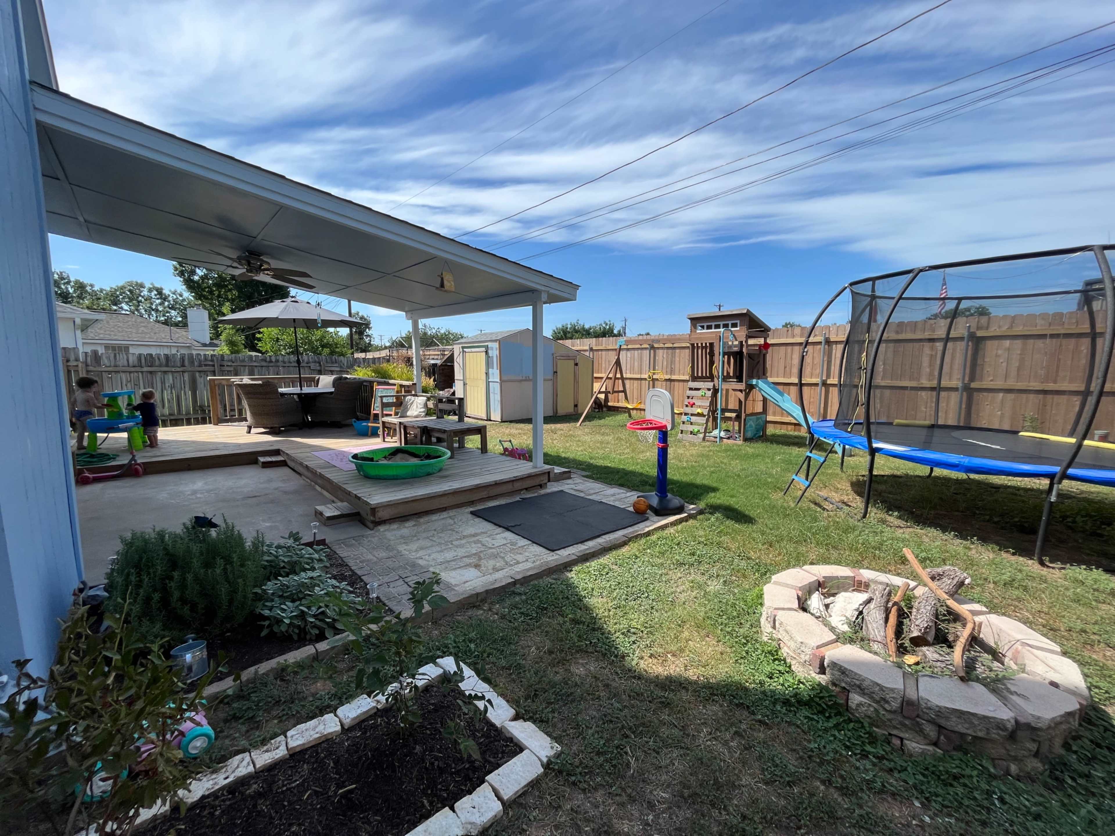 The image shows a backyard with a covered patio, a grassy area, a trampoline, and a playset, surrounded by a wooden fence.