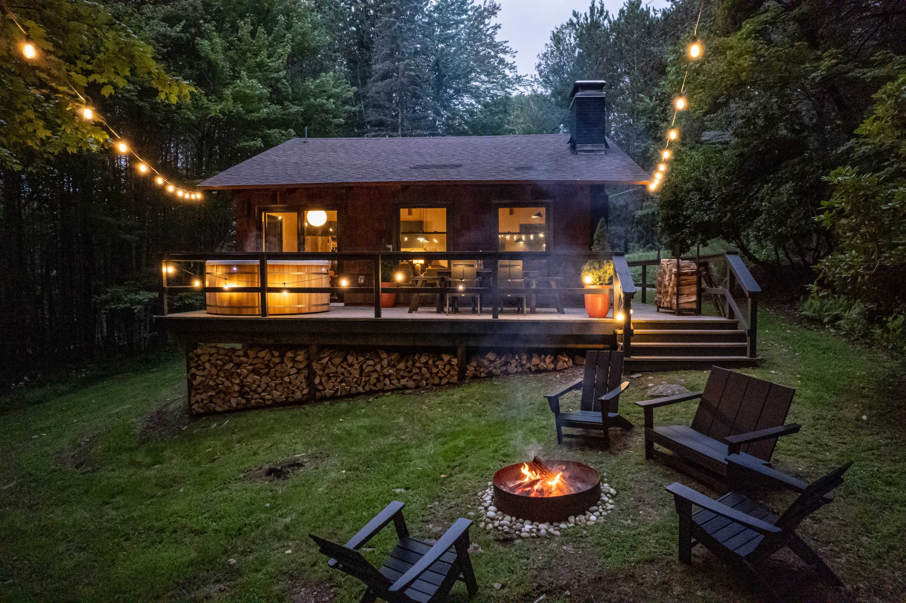 A modern cabin surrounded by trees features a fire pit and string lights illuminating the outdoor seating area.