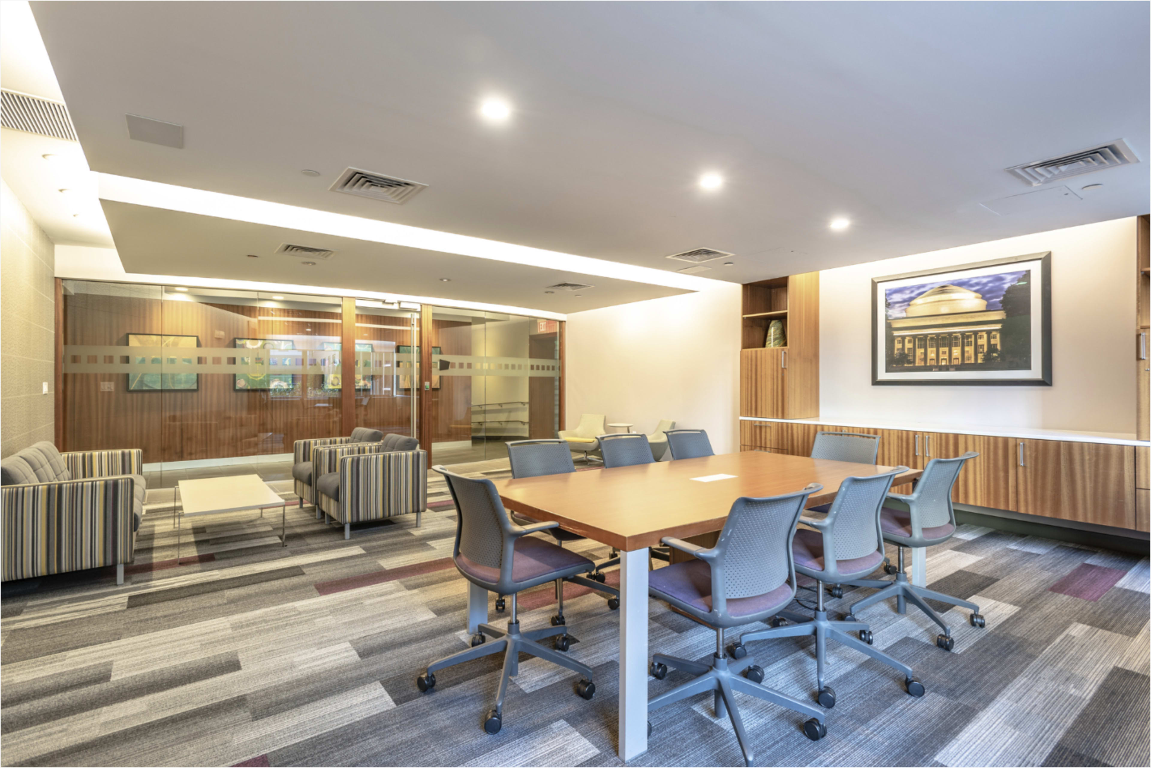 The image shows a modern conference room with a large wooden table surrounded by chairs, accompanied by a glass-walled area that likely serves as a breakout space.
