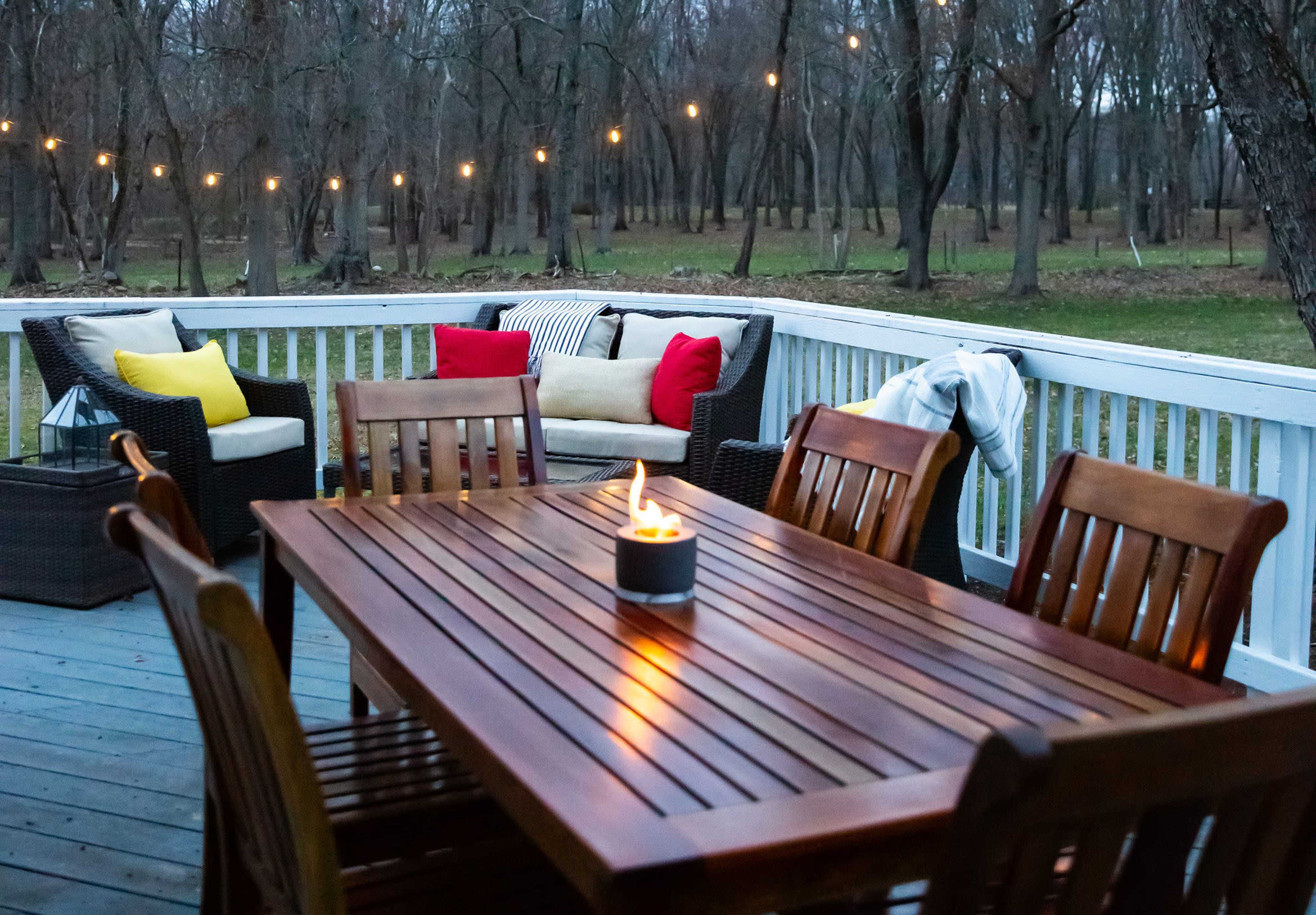 A wooden dining table with chairs sits on a deck, featuring a small flame centerpiece, surrounded by outdoor seating and strung lights in a wooded area.