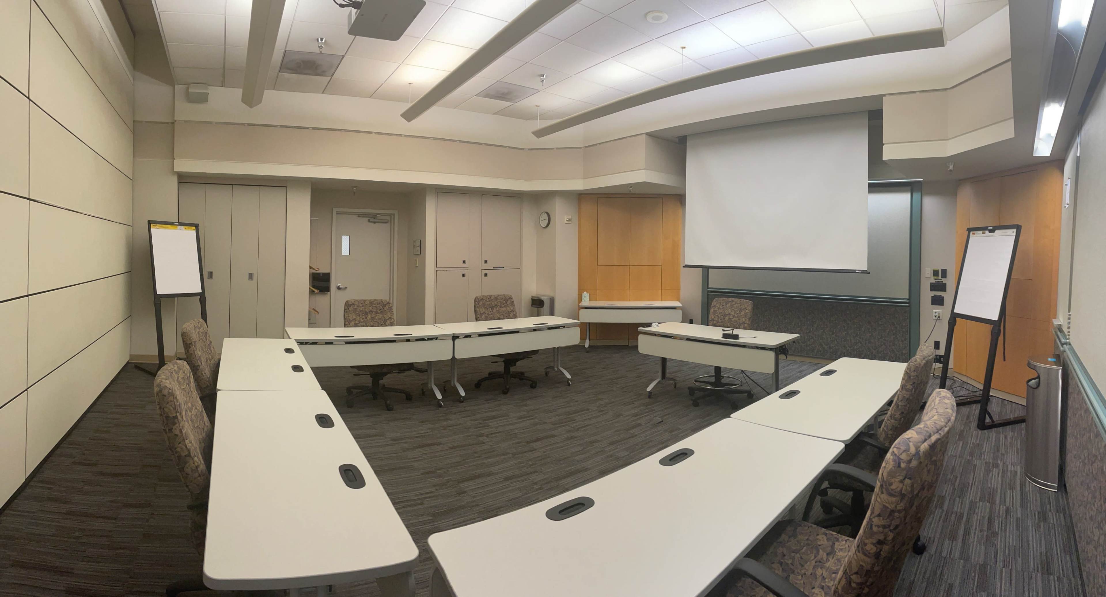 A large conference room features several tables arranged in a U-shape with chairs and a projector screen on one wall.