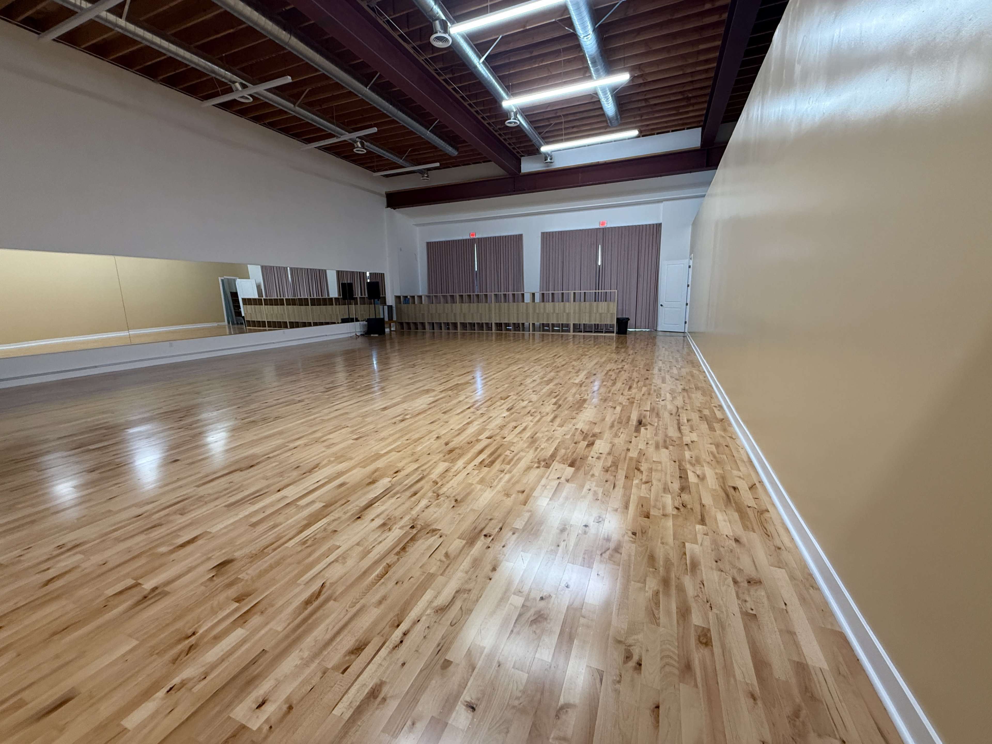 The image shows a spacious dance studio with a polished wooden floor, large mirrors, and beige walls.