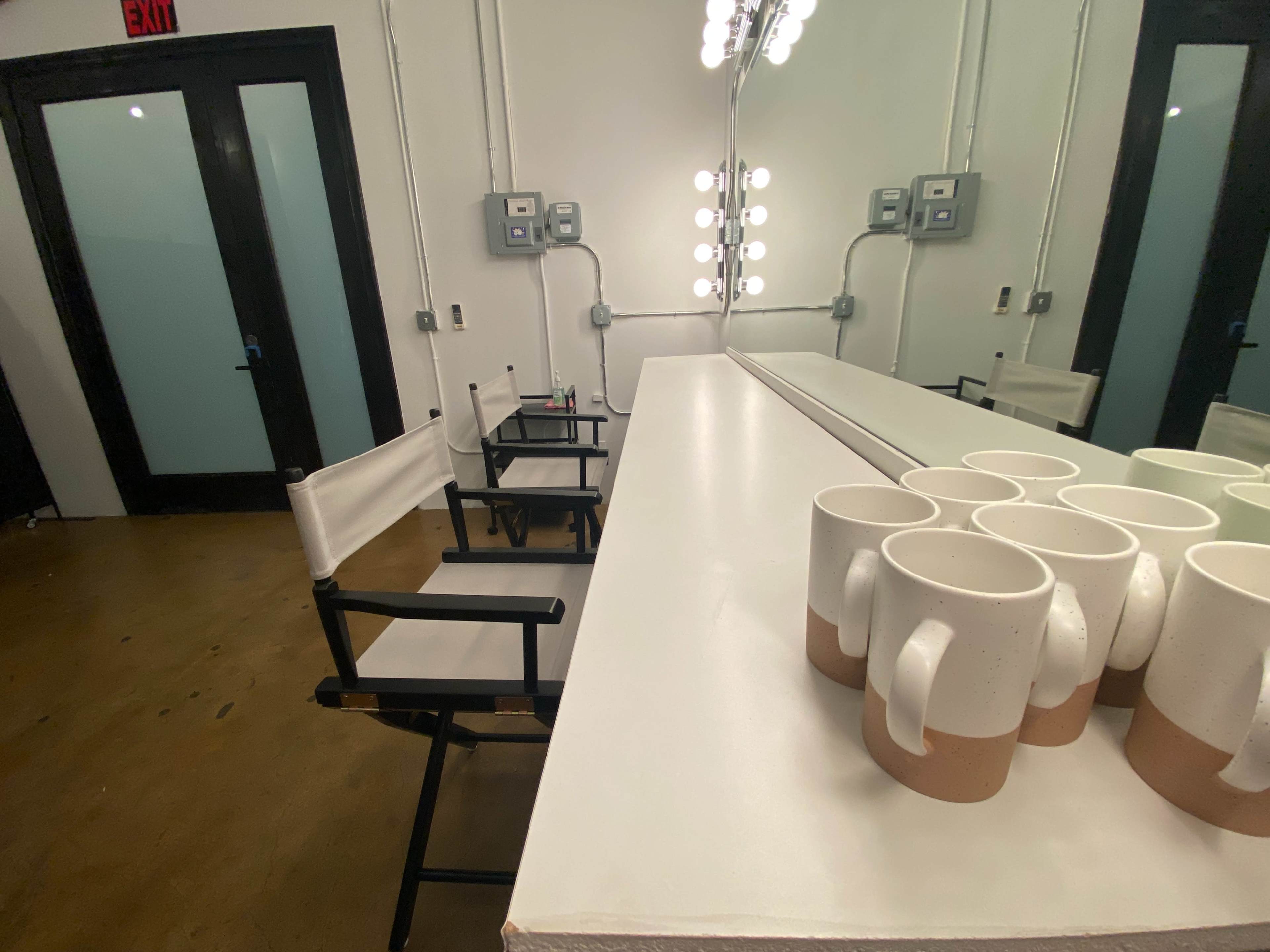 The image shows a brightly lit dressing room with two director's chairs, a long white counter, and a row of ceramic mugs positioned neatly on the counter.