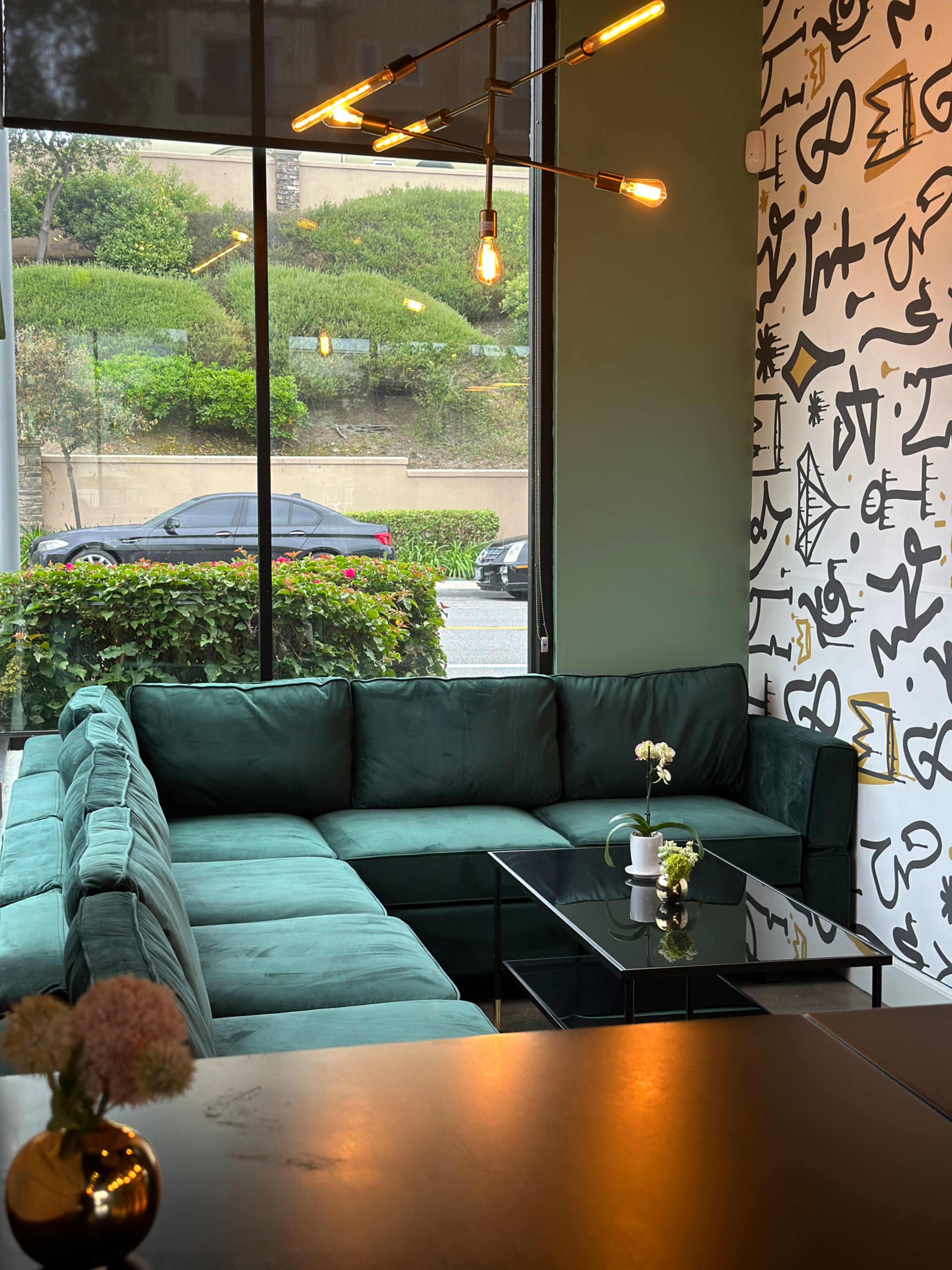 A large green sectional couch is positioned in a well-lit corner of a modern space, next to a glass coffee table and a large window displaying greenery and a street view.