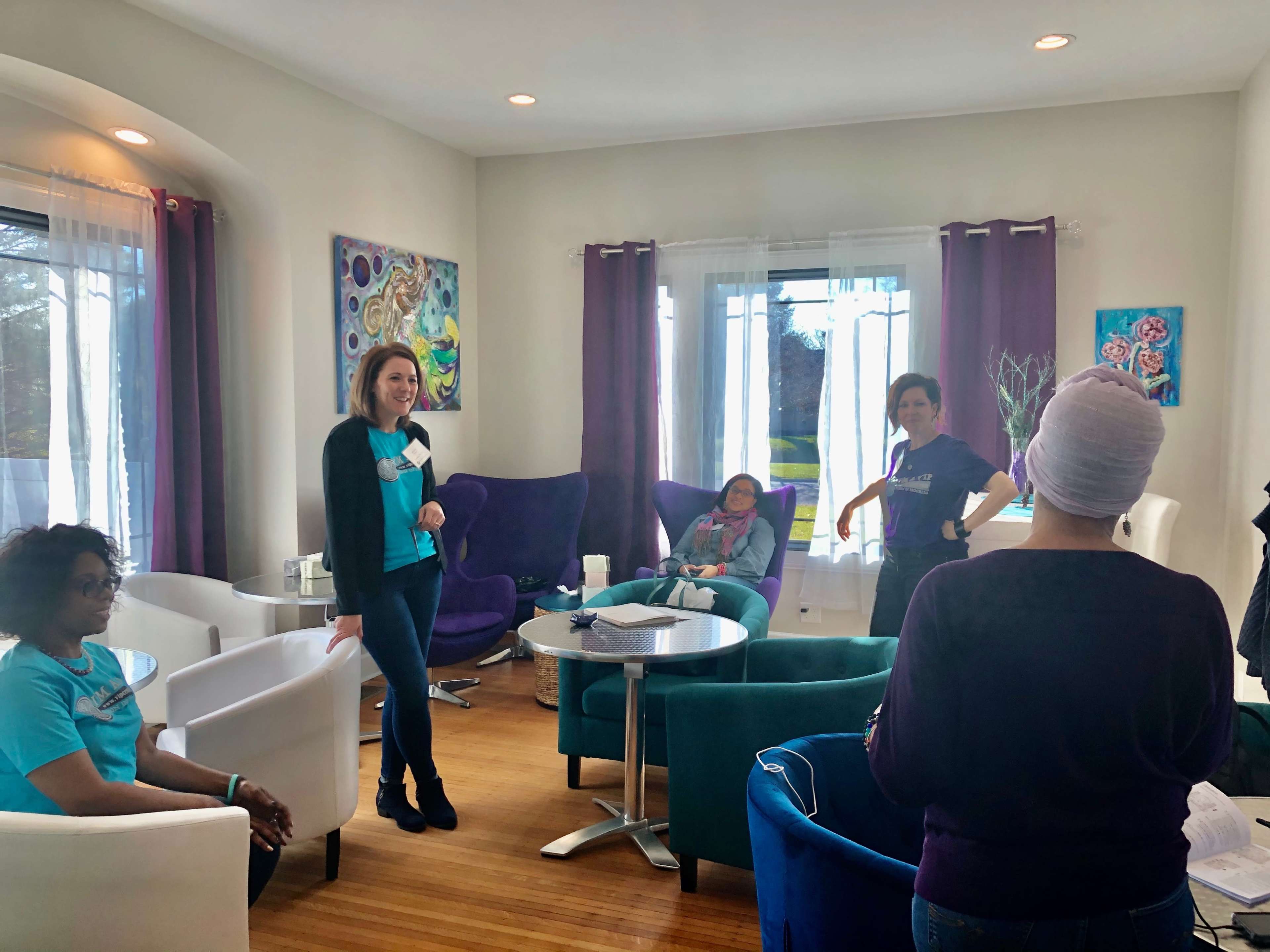 A group of five women engage in conversation in a well-lit room with colorful furniture and artwork on the walls.