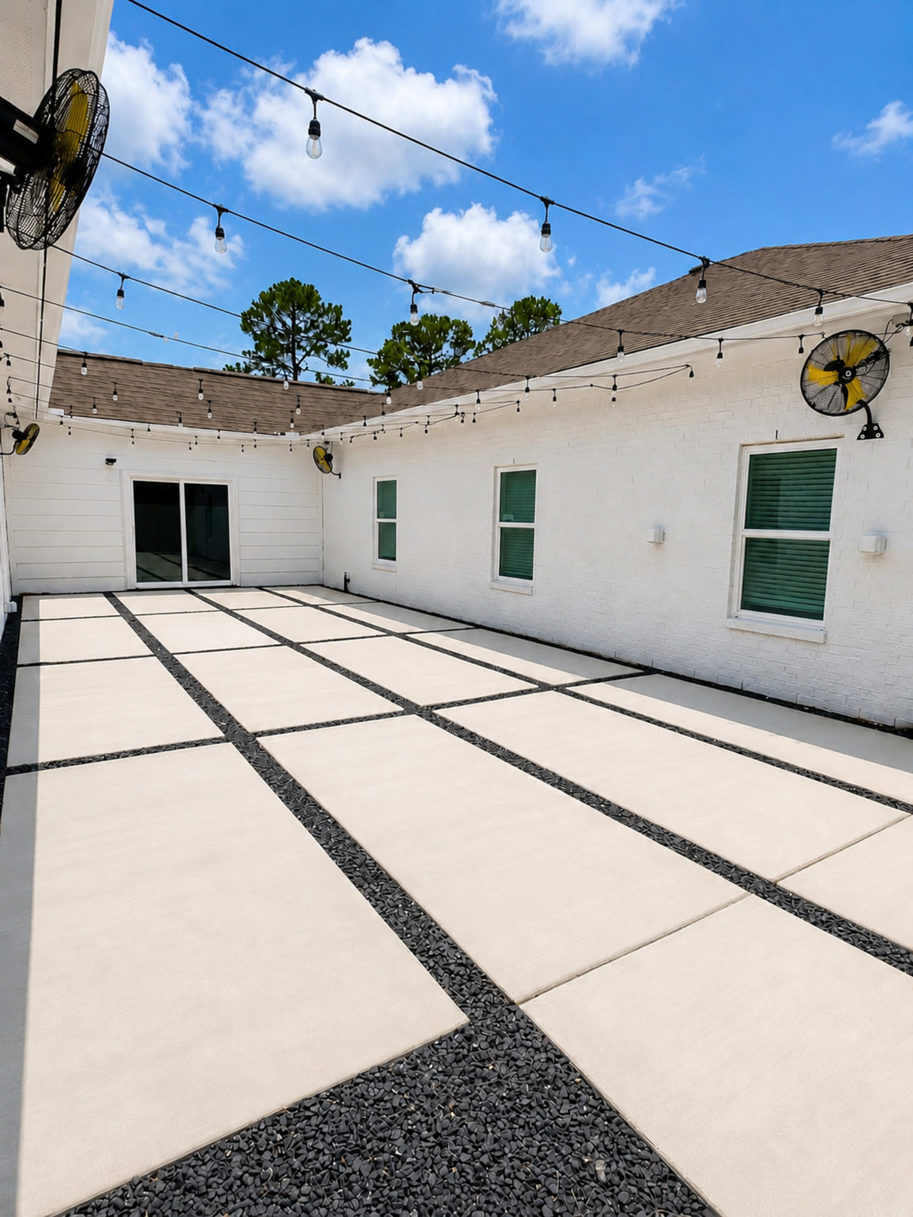 The image shows a spacious outdoor patio featuring large rectangular concrete slabs arranged in a grid over a bed of small gravel, with string lights and wall-mounted fans overhead.