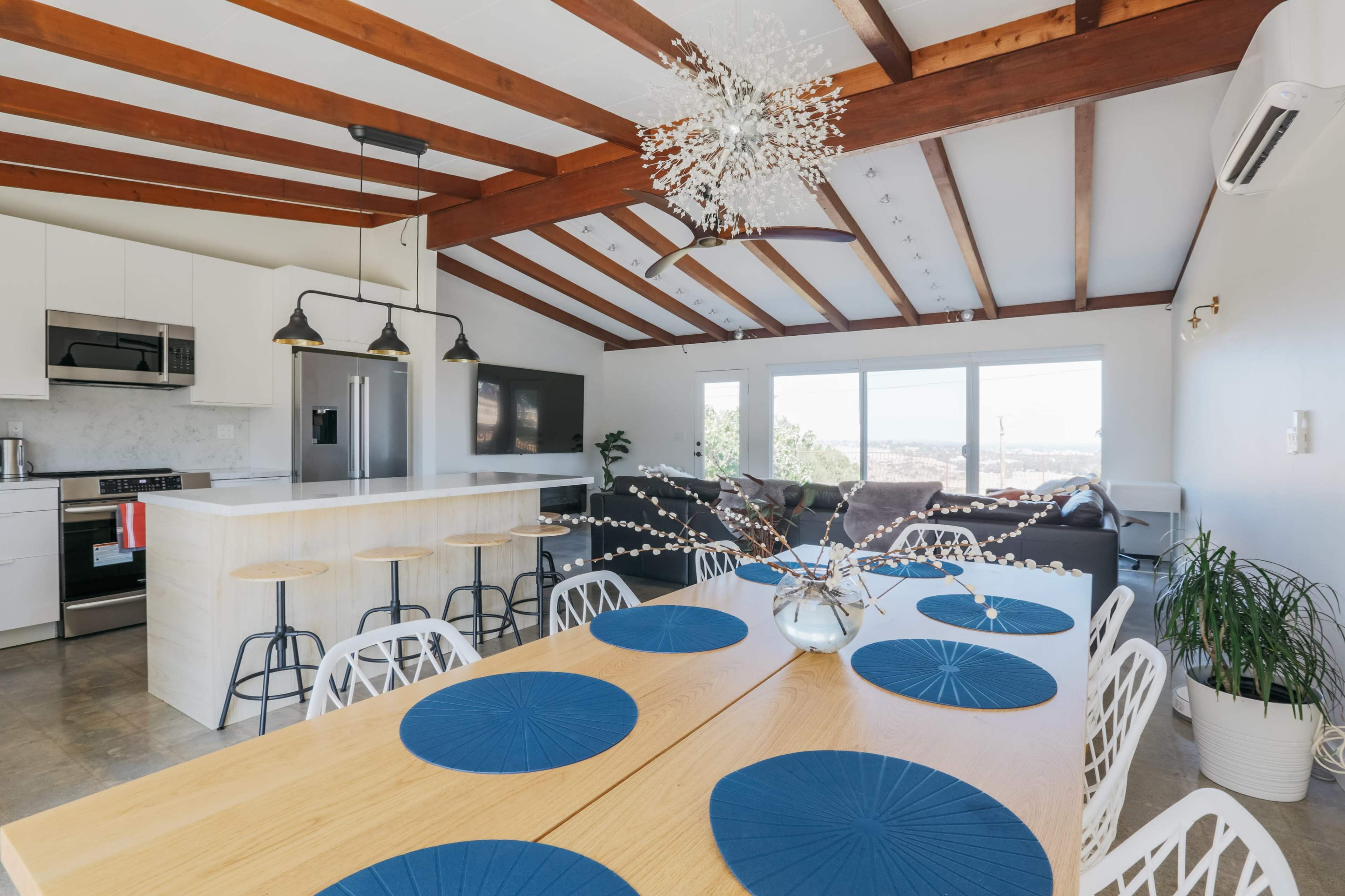 A modern open-concept kitchen and dining area features wooden beams, a spacious island, and a large dining table set with blue placemats.
