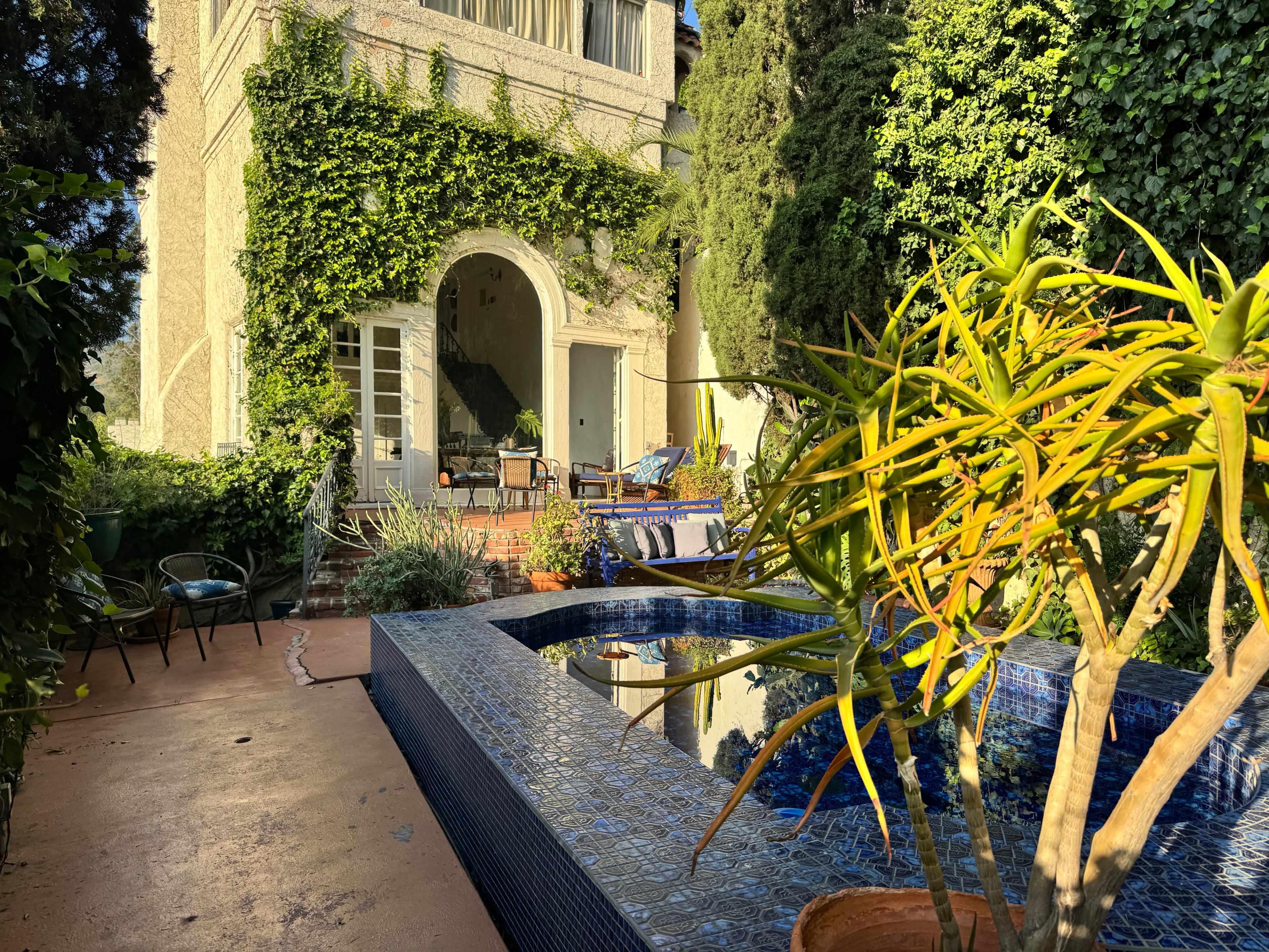 A house surrounded by greenery features a blue-tiled swimming pool and various outdoor seating areas.