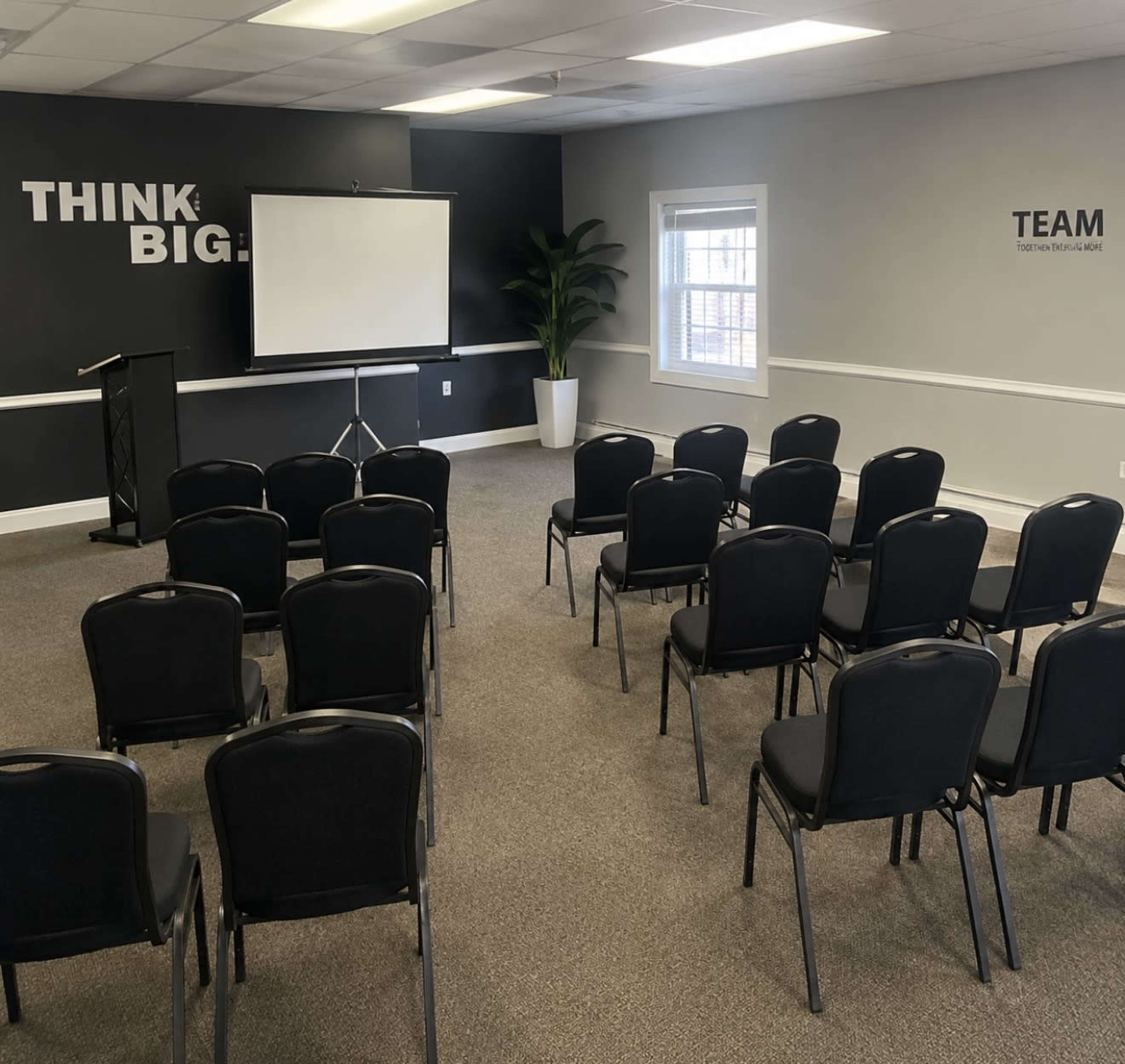The image shows a room set up for a presentation, featuring a projector screen at the front and rows of black chairs arranged facing it.