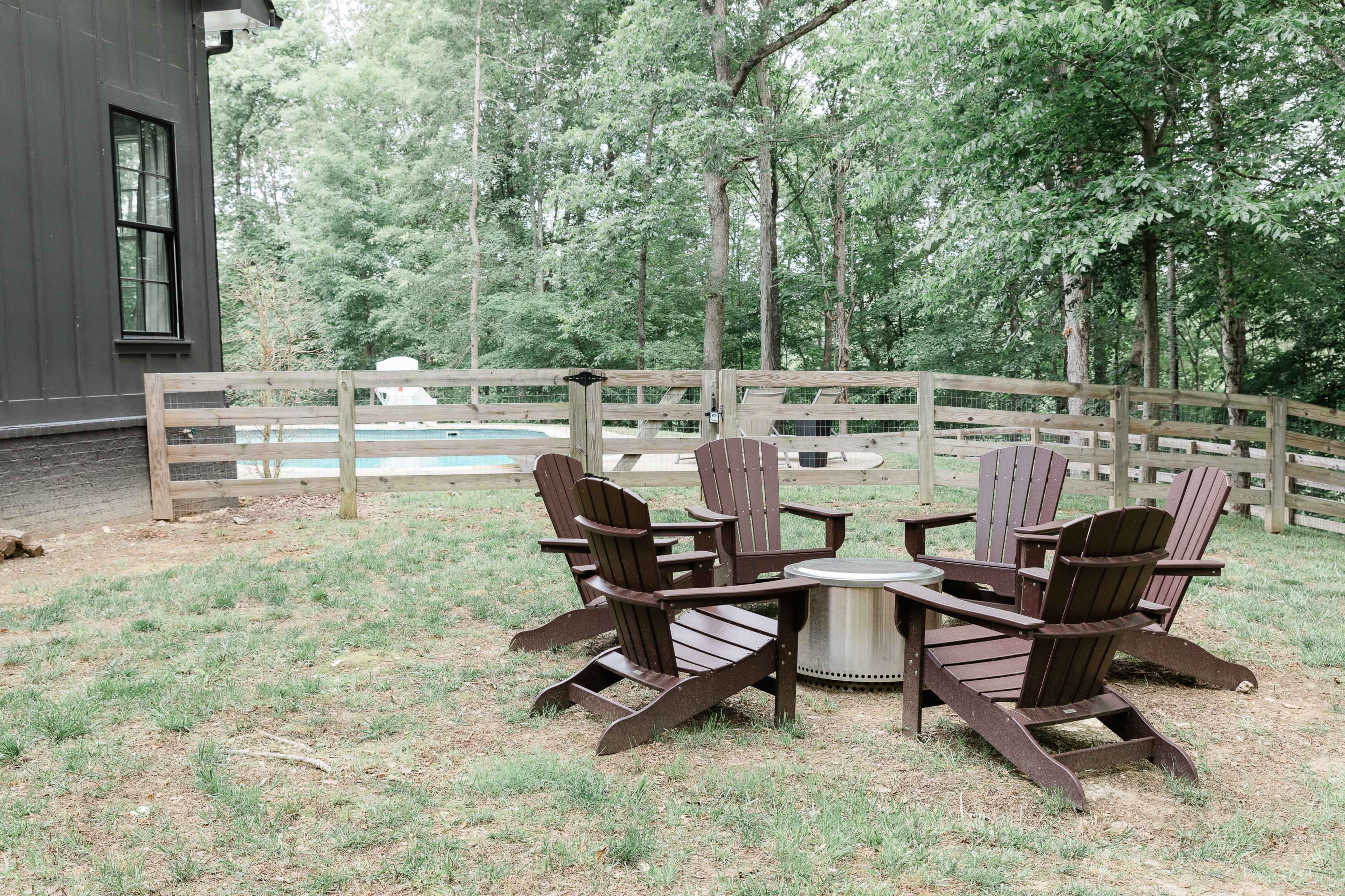 A set of four wooden Adirondack chairs surrounds a fire pit in a grassy area next to a fenced yard with a pool.