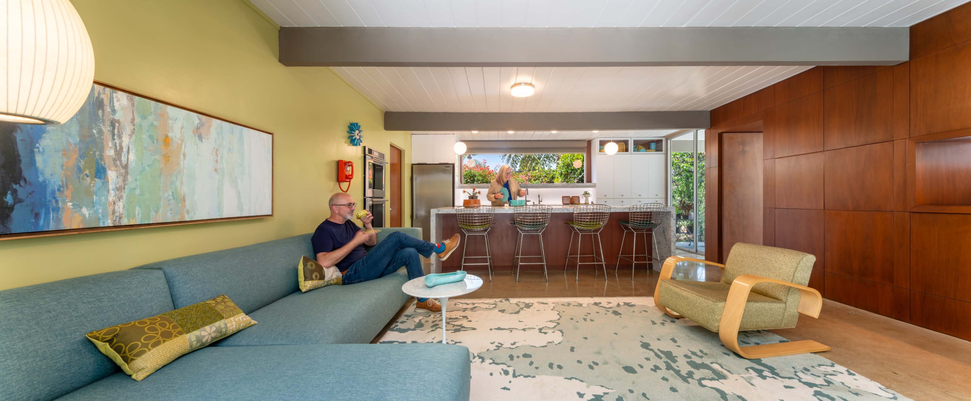 A modern living room features a blue couch, a decorative rug, and a kitchen visible in the background with bar stools and a person preparing food.