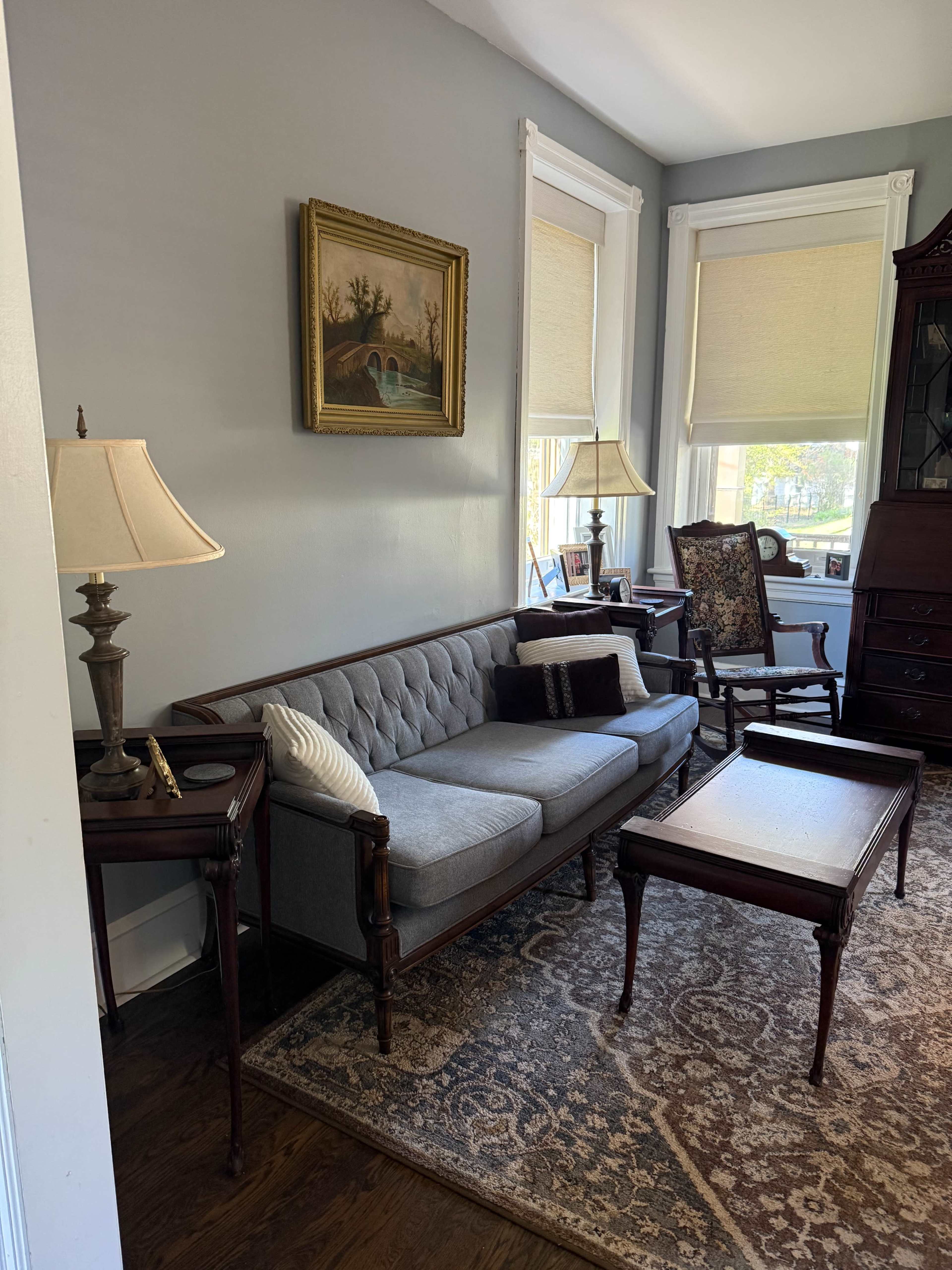 The image shows a living room with a gray upholstered sofa, a wooden coffee table, two lamps, and a framed painting on the wall.
